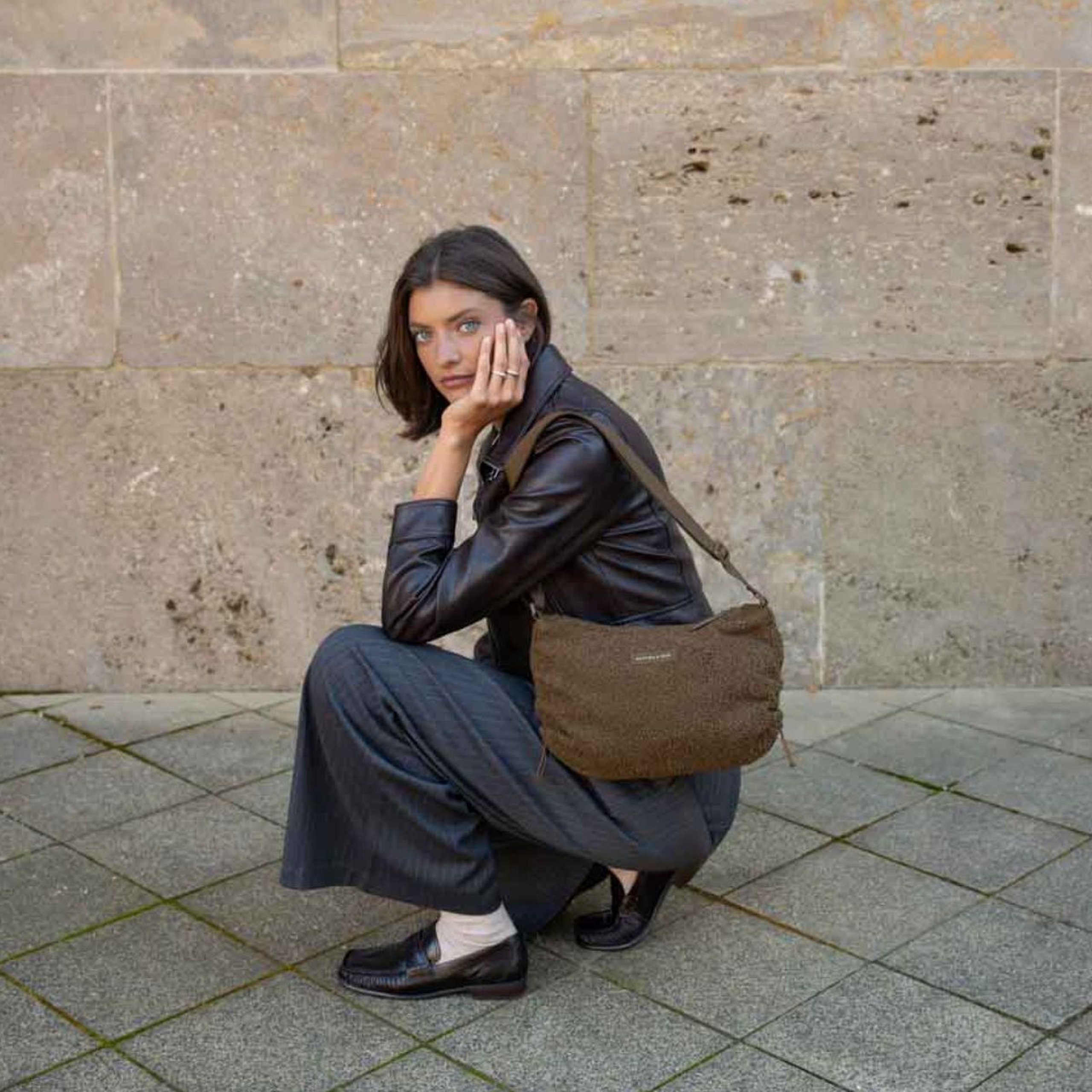 Frau in Lederjacke mit einer Kapten & Son Schultertasche aus weichem Fleece, die vor einer Steinwand in der Hocke sitzt.