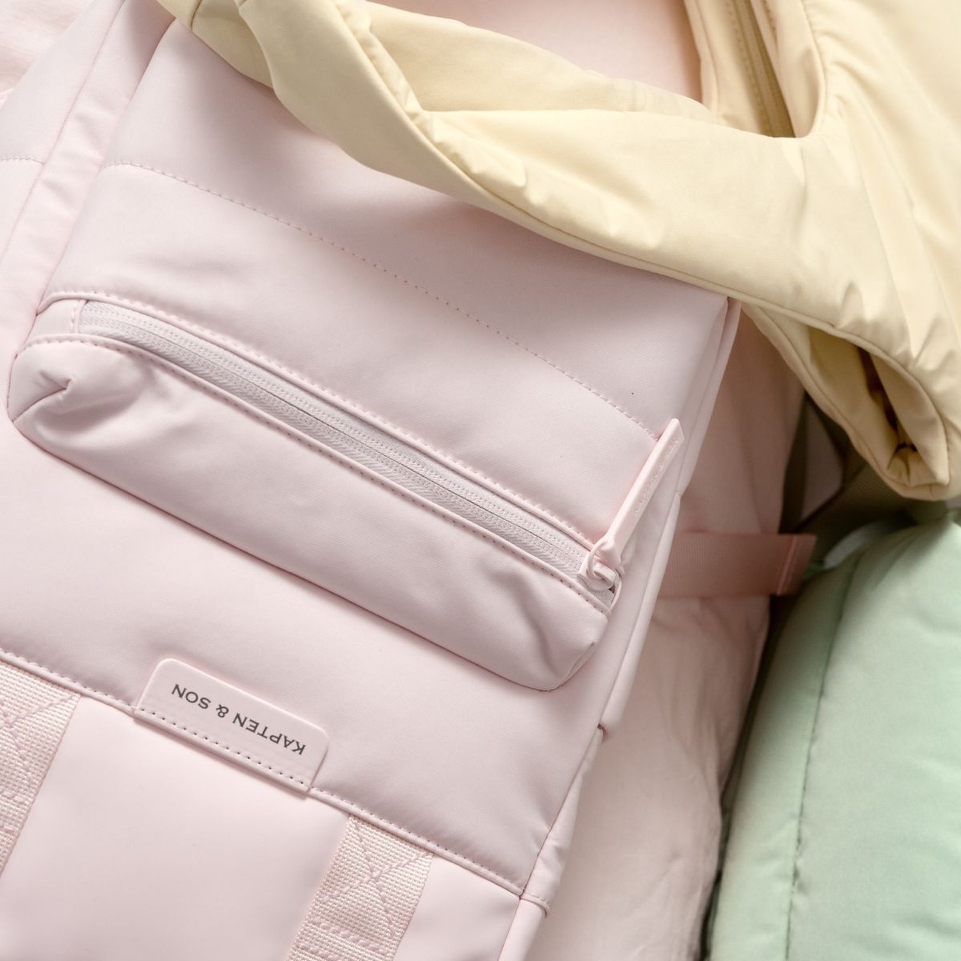 Nahaufnahme eines pastellrosa Rucksacks mit sichtbarer Reißverschlusstasche und beigen Trägern. Er liegt neben weichem Stoff in Creme- und Mintgrüntönen.