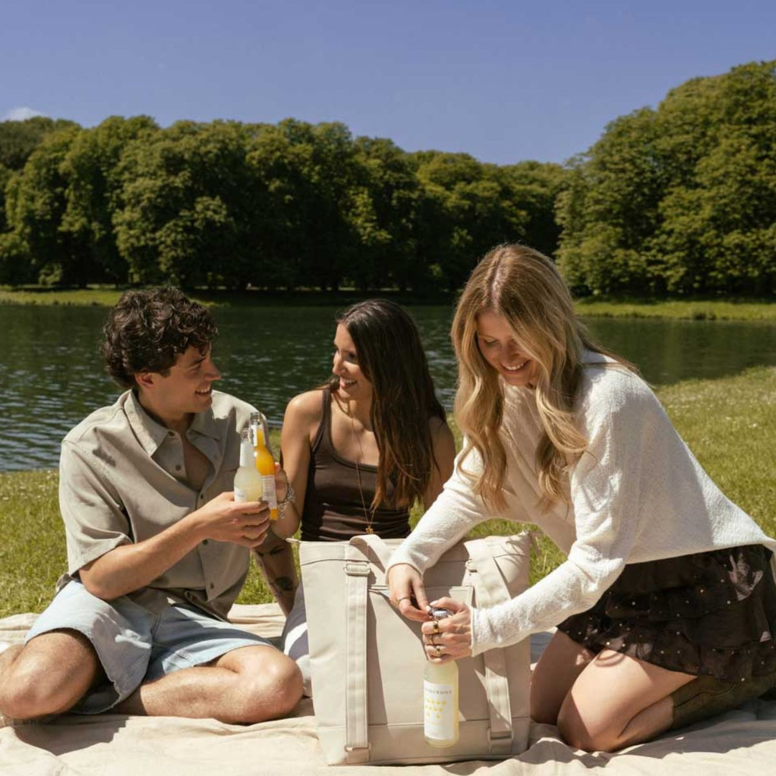 Three people enjoy a sunny picnic by a lake, sitting on a blanket. They smile, holding bottles, with trees and a bright blue sky in the background.
