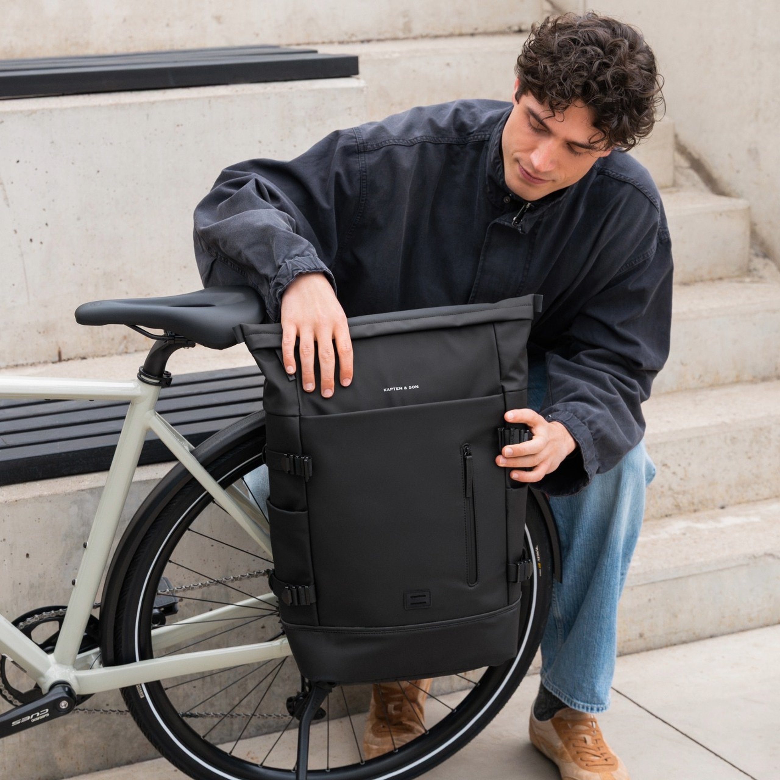 junger mann befestigt schwarzen kapten & son commuter rucksack am gepäckträger seines fahrrads