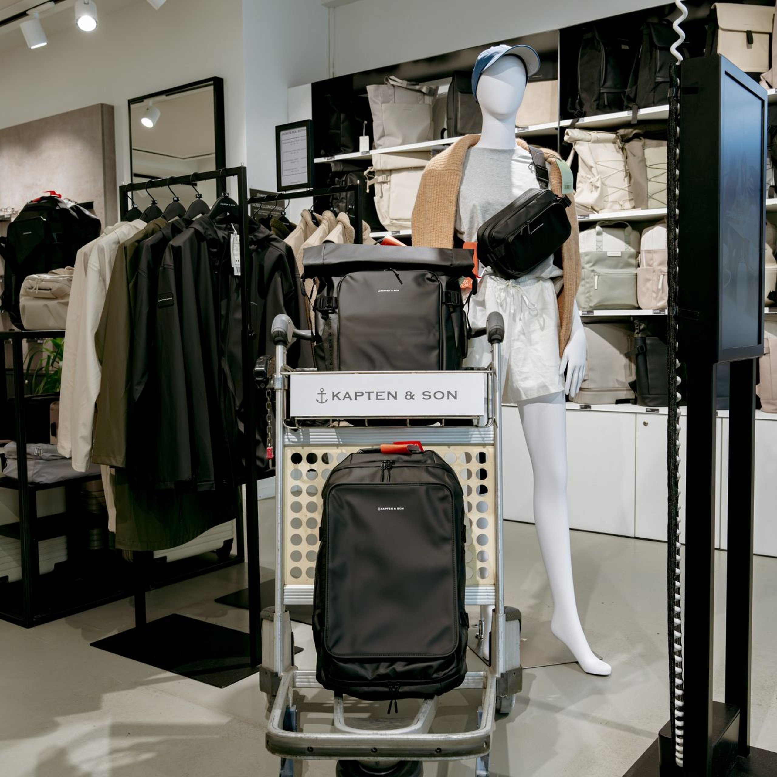 Vue intérieure d'un magasin Kapten & Son avec un chariot à bagages et un mannequin. Le chariot présente des sacs à dos noirs de Kapten & Son, tandis que le mannequin porte une banane noire de Kapten & Son.