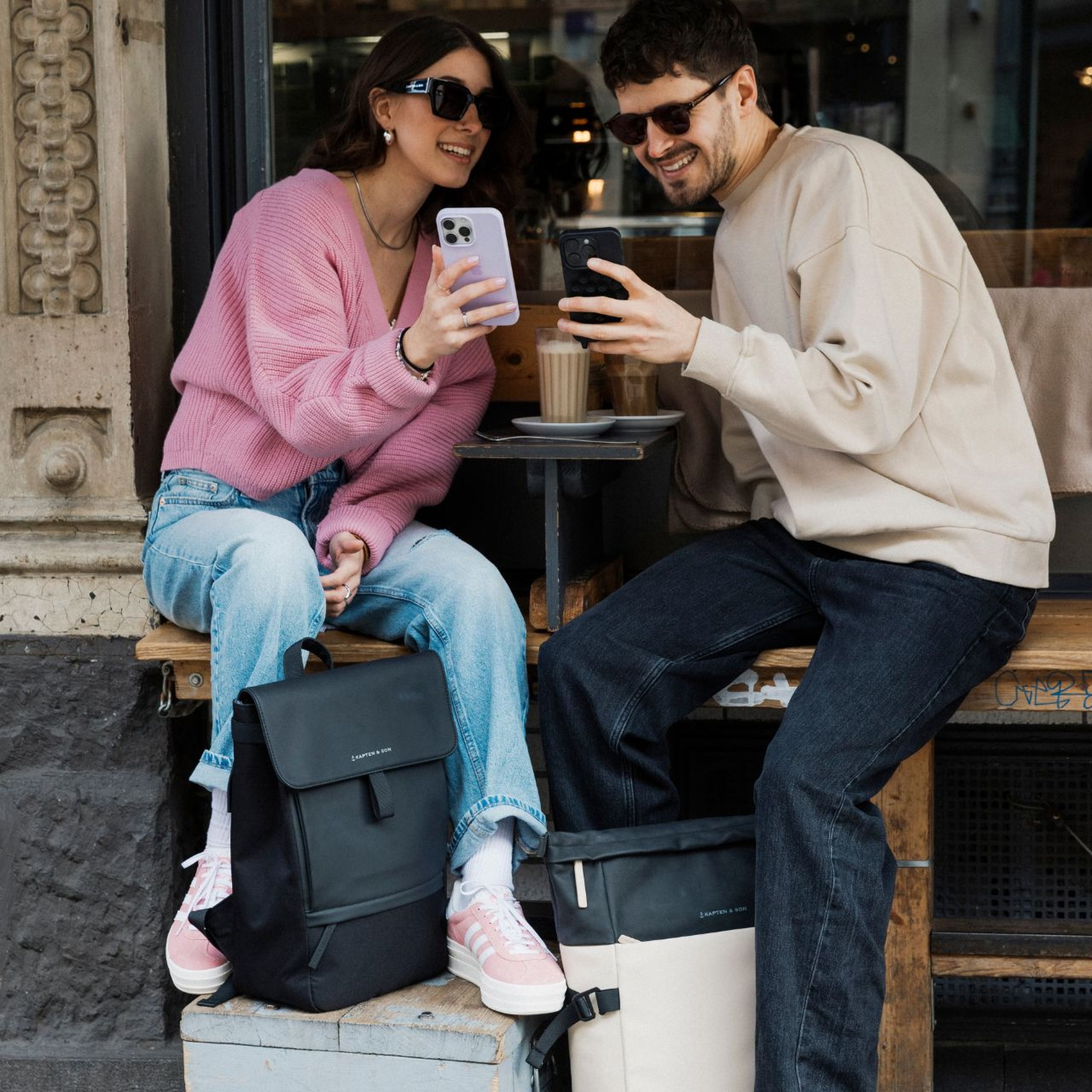 Une femme et un homme souriants assis devant un café, regardant ensemble leurs smartphones. Tous deux portent des lunettes de soleil de Kapten & Son. Devant eux se trouvent un sac à dos noir et un sac à dos beige clair de Kapten & Son.