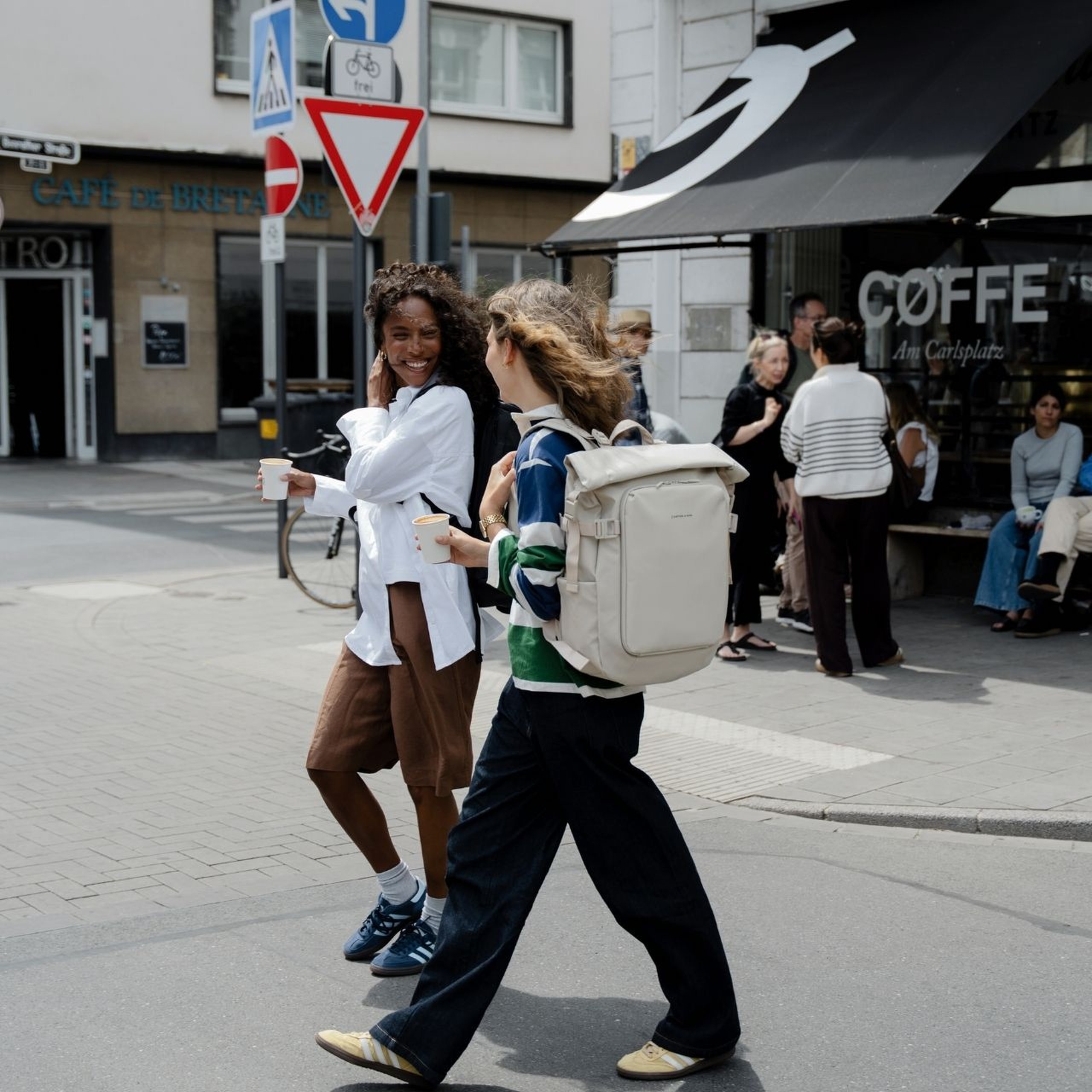 Twee vrouwen lachen terwijl ze een straat in de stad oversteken. Een van hen draagt een crèmekleurige Kapten & Son rugzak.