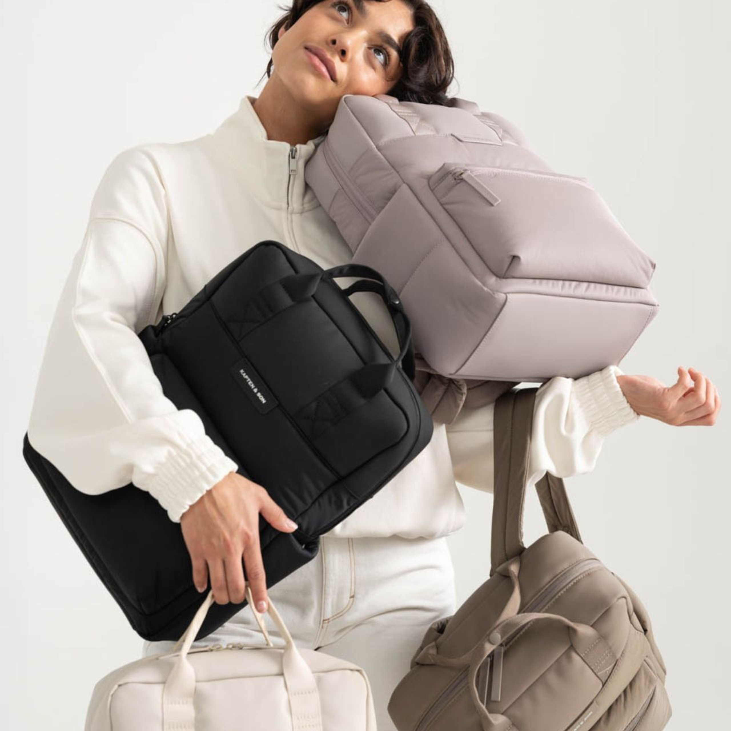 Eine Frau in einem weißen Outfit jongliert auf humorvolle Weise mit vier Taschen, darunter eine schwarze und eine beige. Sie blickt nach oben und strahlt Leichtigkeit und Verspieltheit aus.