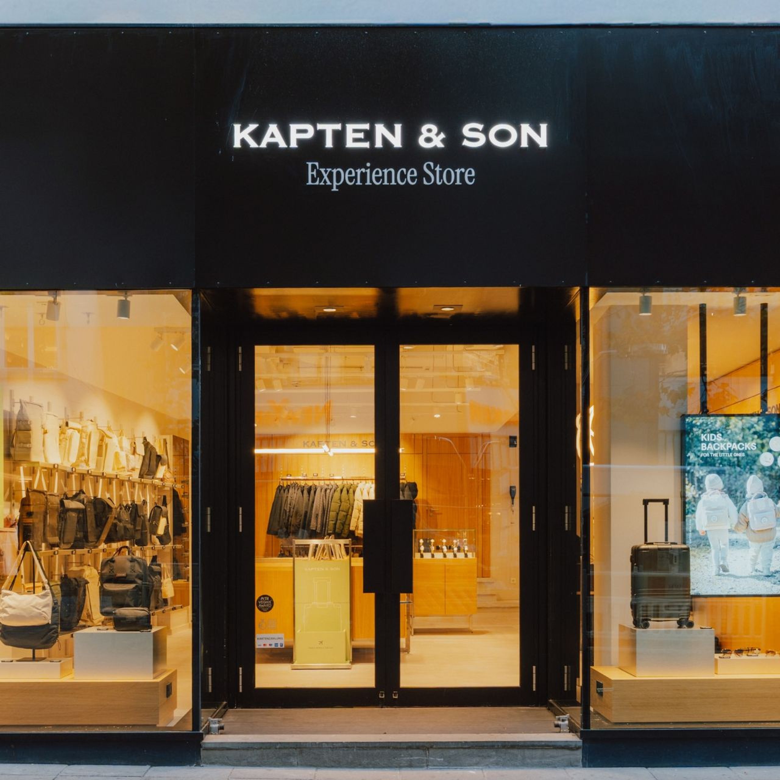 Kapten & Son Experience Store – Ladenfront mit beleuchtetem Logo, Rucksäcken, Taschen und Reisegepäck in den Schaufenstern