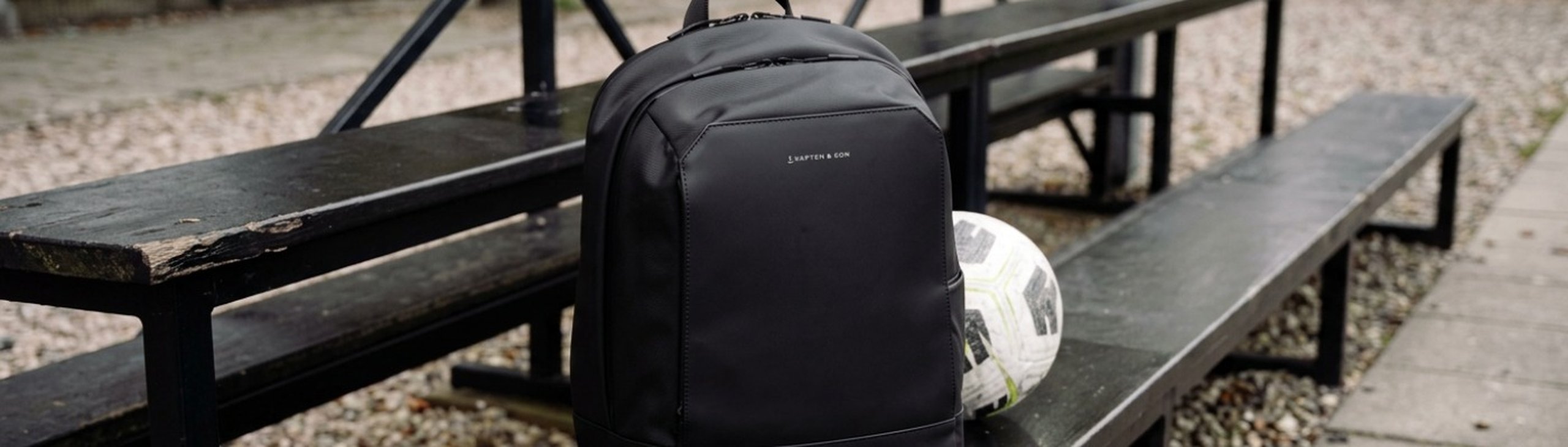 schwarzer rucksack von kapten and son steht auf einer schwarzen bierbank neben einem fußball