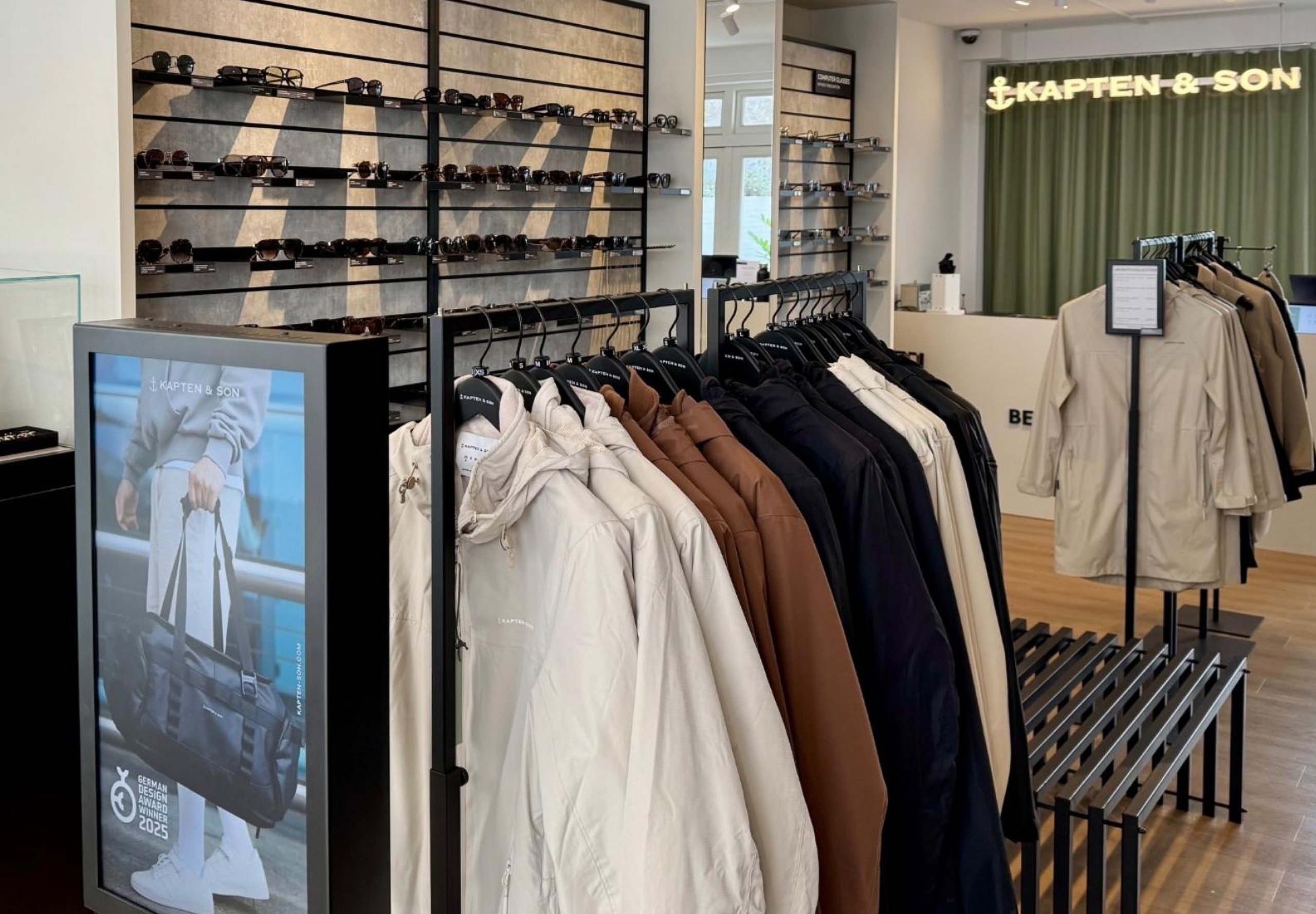 Un intérieur de boutique élégant avec un portant de vestes dans des tons neutres et un mur orné de rangées de lunettes de soleil. L'enseigne Kapten & Son est illuminée en arrière-plan.