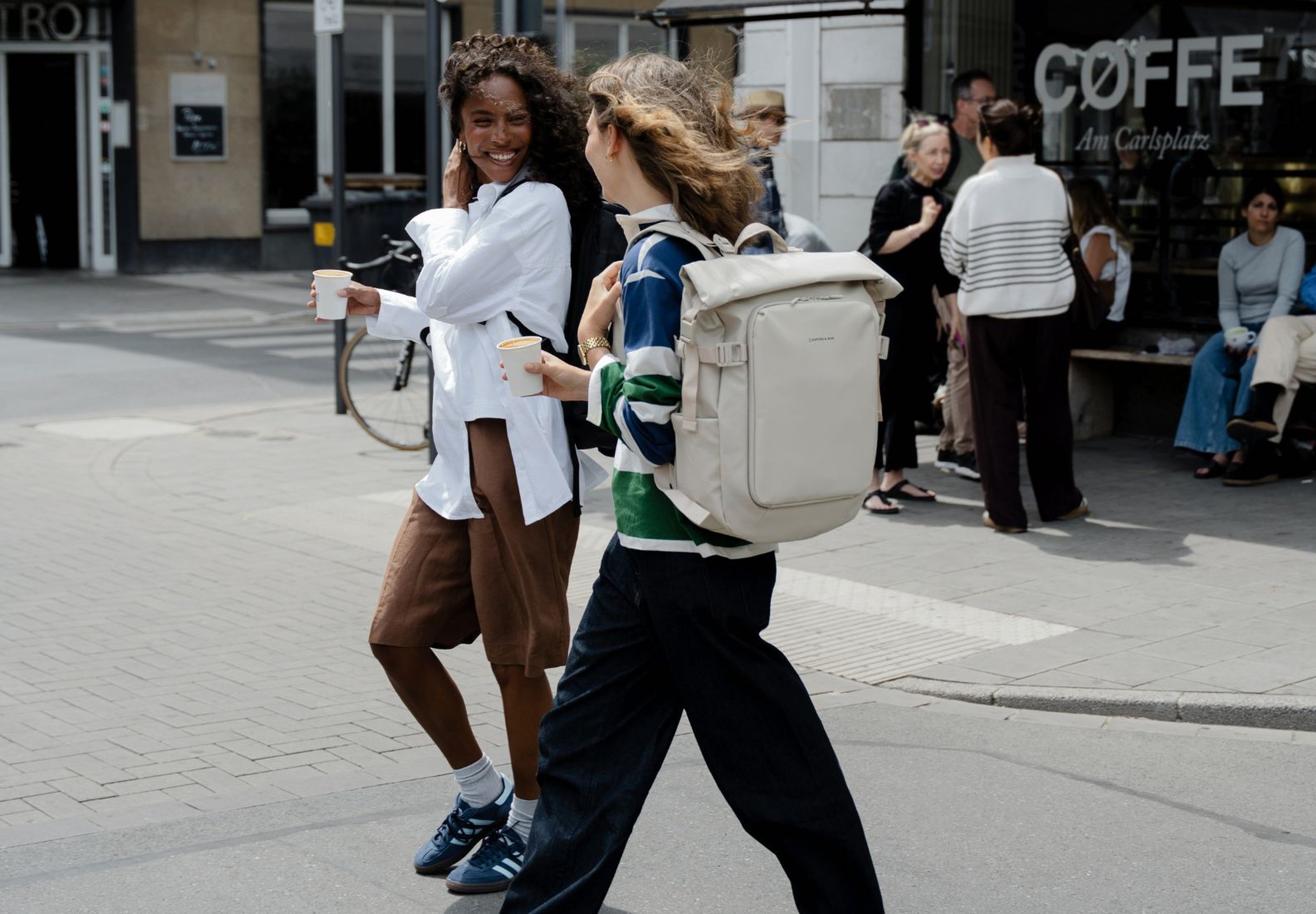 Deux femmes avec des sacs à dos se promènent et discutent joyeusement près d'un café dans une rue de la ville. Des personnes se rassemblent devant le café, créant une atmosphère animée.