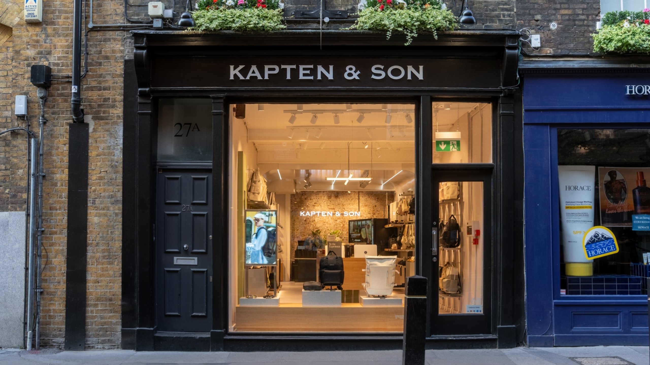Kapten & Son Experience Store – Ladenfront mit beleuchtetem Logo, Rucksäcken, Taschen und Reisegepäck in den Schaufenstern