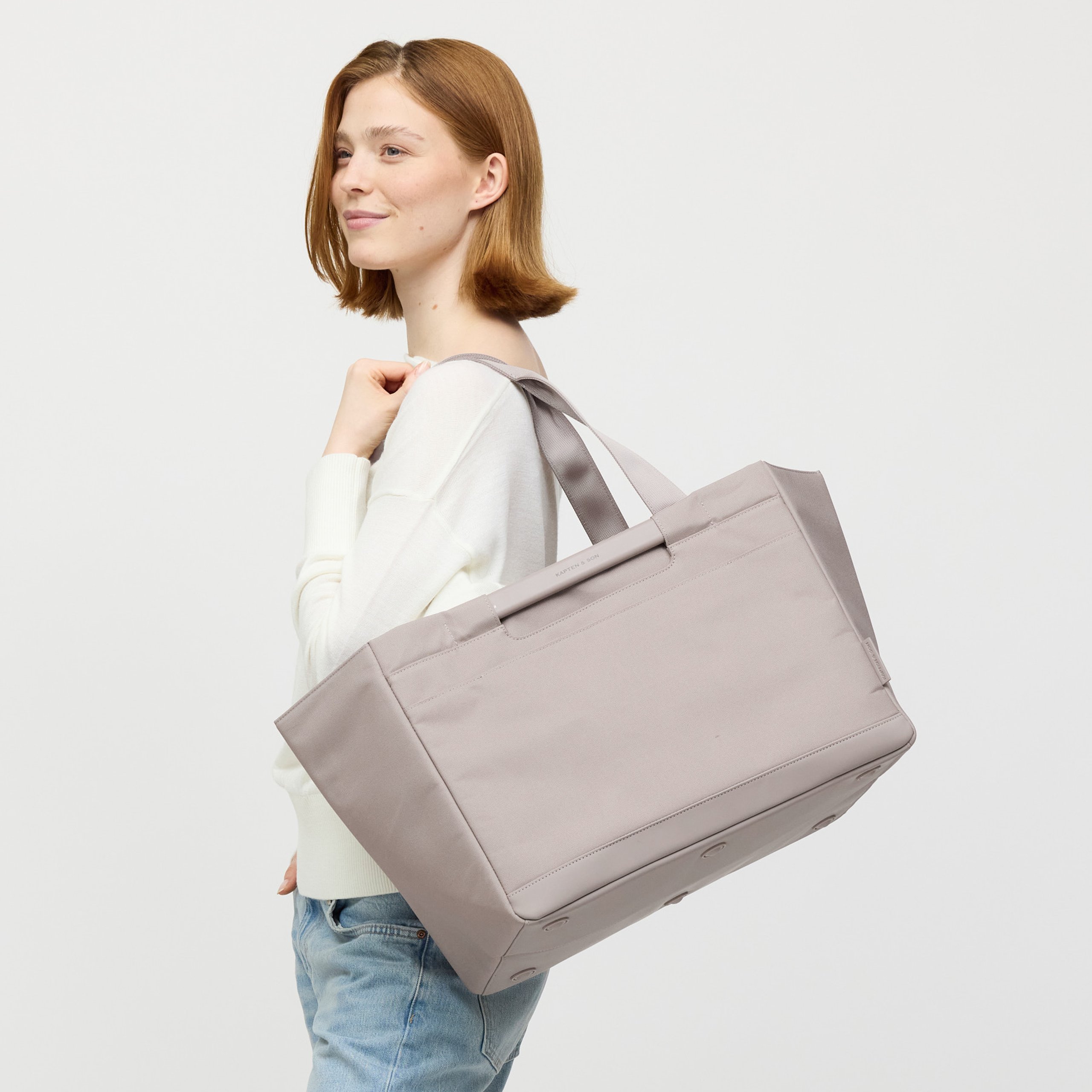 Eine lächelnde Frau trägt eine große olivgrüne Tote Bag mit Struktur über der rechten Schulter. Sie trägt ein weißes Langarmshirt und blaue Jeans. Die Tasche hat einen gerippten Stoffkörper und einen glatten Boden. Der Hintergrund ist hellgrau.