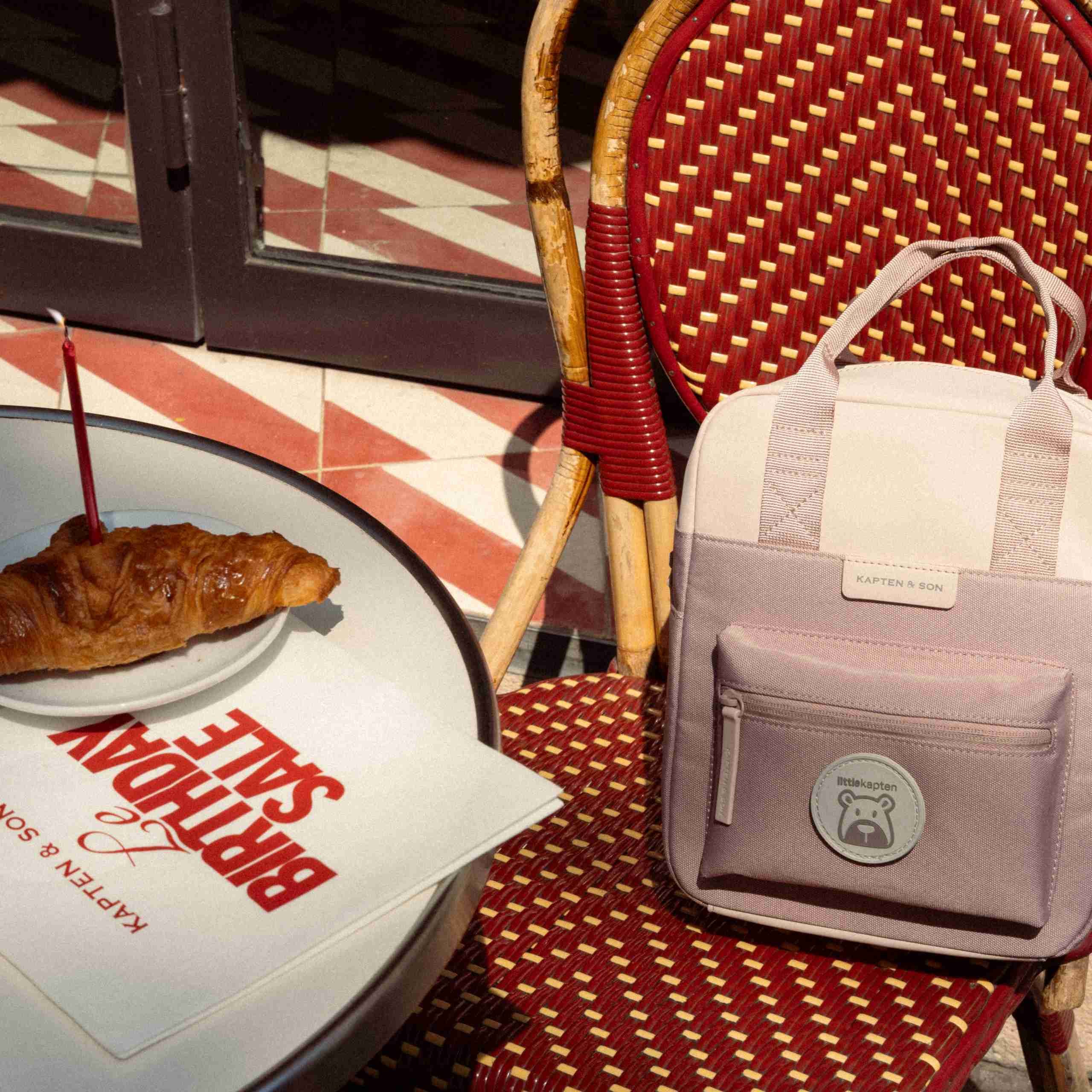 Ein kleiner zweifarbiger Kinderrucksack in Creme und Mauve von Kapten & Son mit einem „littlekapten" Bären-Patch, auf einem roten Korbstuhl eines Cafés neben einem Bistrotisch mit einem Croissant und einer Geburtstagskerze auf einem Teller sowie einem „Le Birthday Sale – Kapten & Son" Flyer, auf einem gemusterten Fliesenboden.