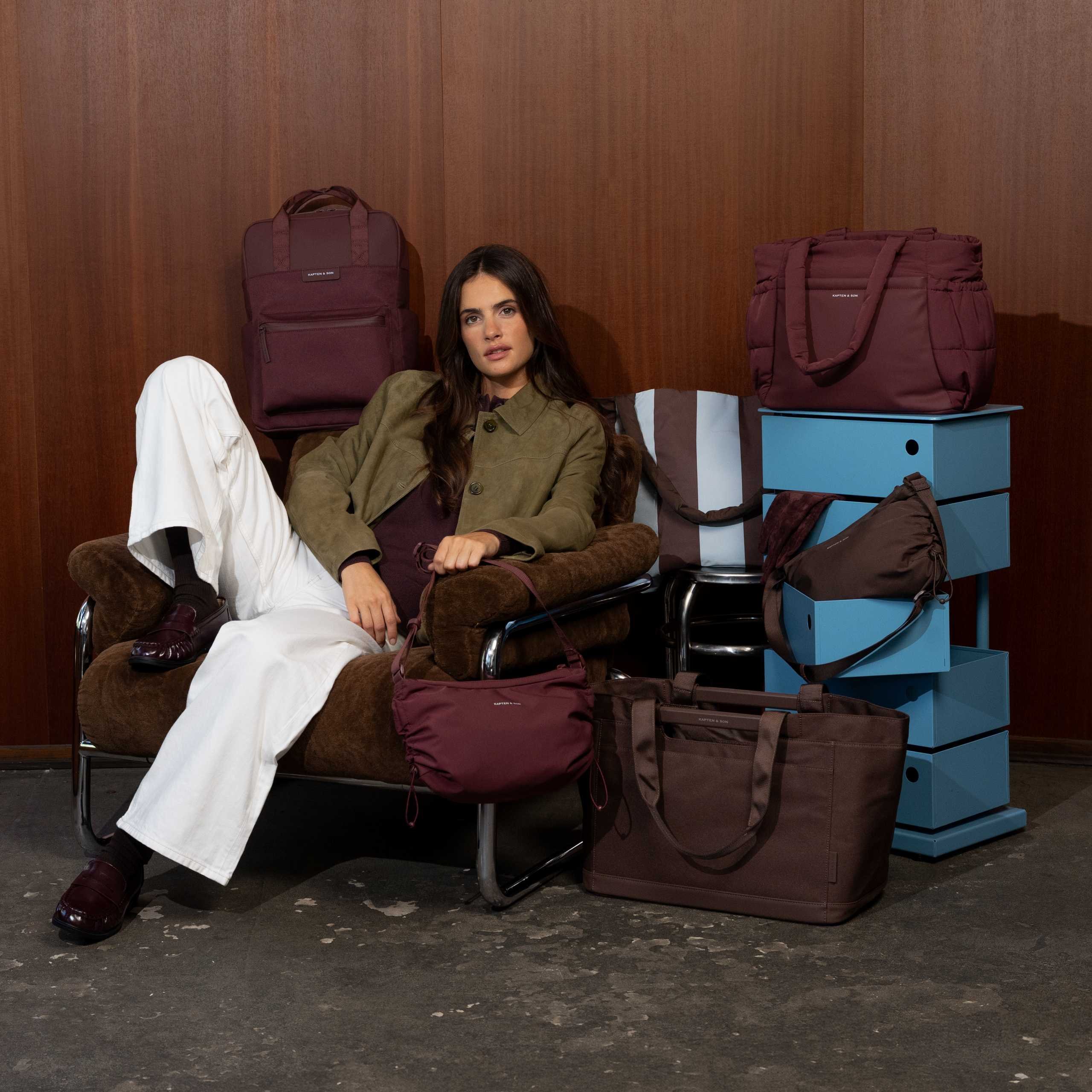 Jeune femme aux longs cheveux foncés assise de façon détendue dans un fauteuil en velours marron, portant une veste en daim vert olive, un pantalon large blanc et des mocassins bordeaux, entourée de plusieurs sacs Kapten & Son en bordeaux et marron chocolat : un sac à dos Bergen bordeaux, un grand sac tote matelassé Hellvi Cloud bordeaux, un sac à bandoulière cloud bordeaux sur ses genoux, un sac tote Vaasa marron chocolat au sol et un sac bandoulière marron foncé à cordon sur une étagère bleue, devant un mur lambrissé en bois foncé.