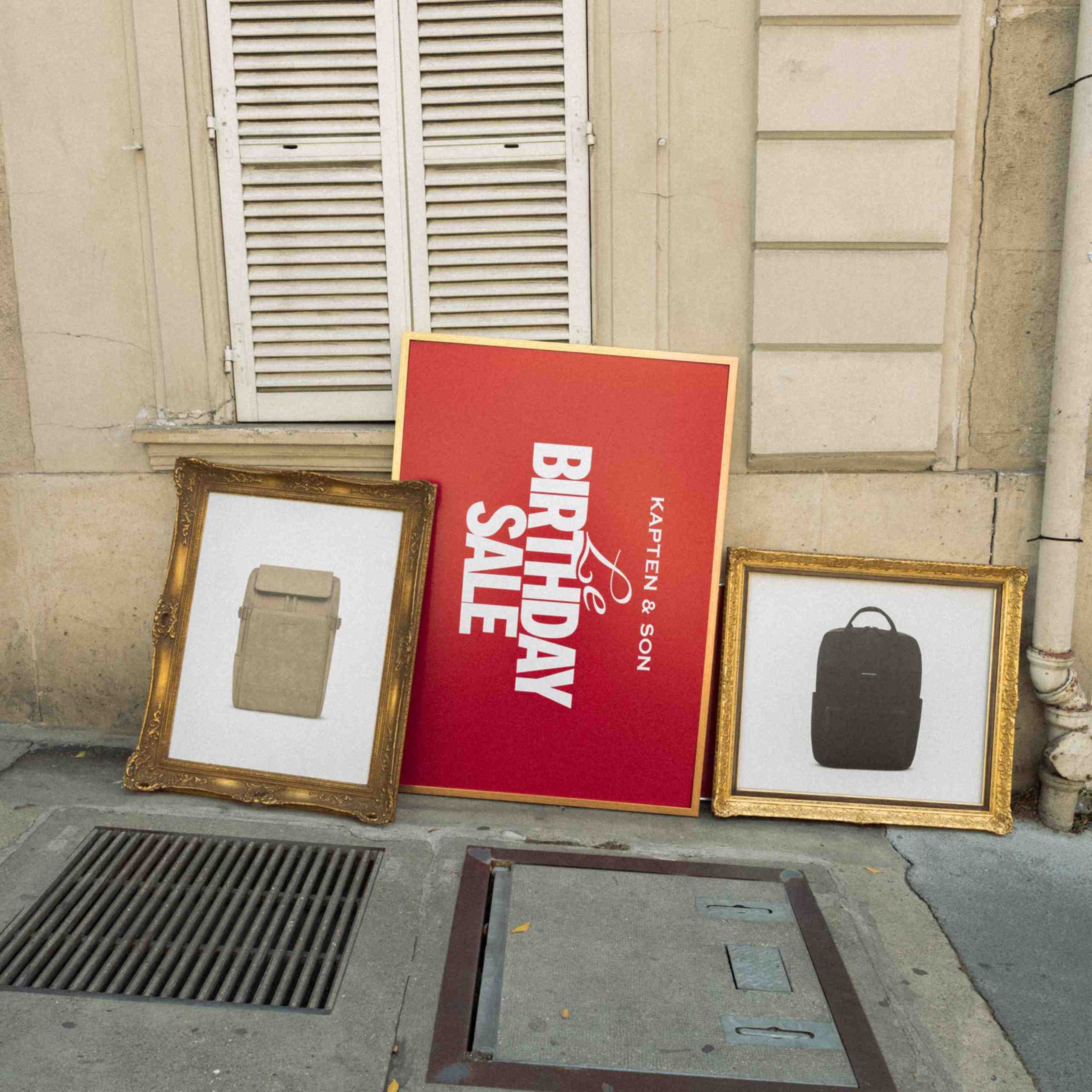 Drei gerahmte Bilder lehnen an einer Pariser Hausfassade: ein goldener Zierrahmen mit einem beigefarbenen Kapten & Son Rucksack, ein rotes Poster in einem Goldrahmen mit der Aufschrift „Le Birthday Sale – Kapten & Son" und ein zweiter Goldrahmen mit einem dunkelgrauen Kapten & Son Rucksack.