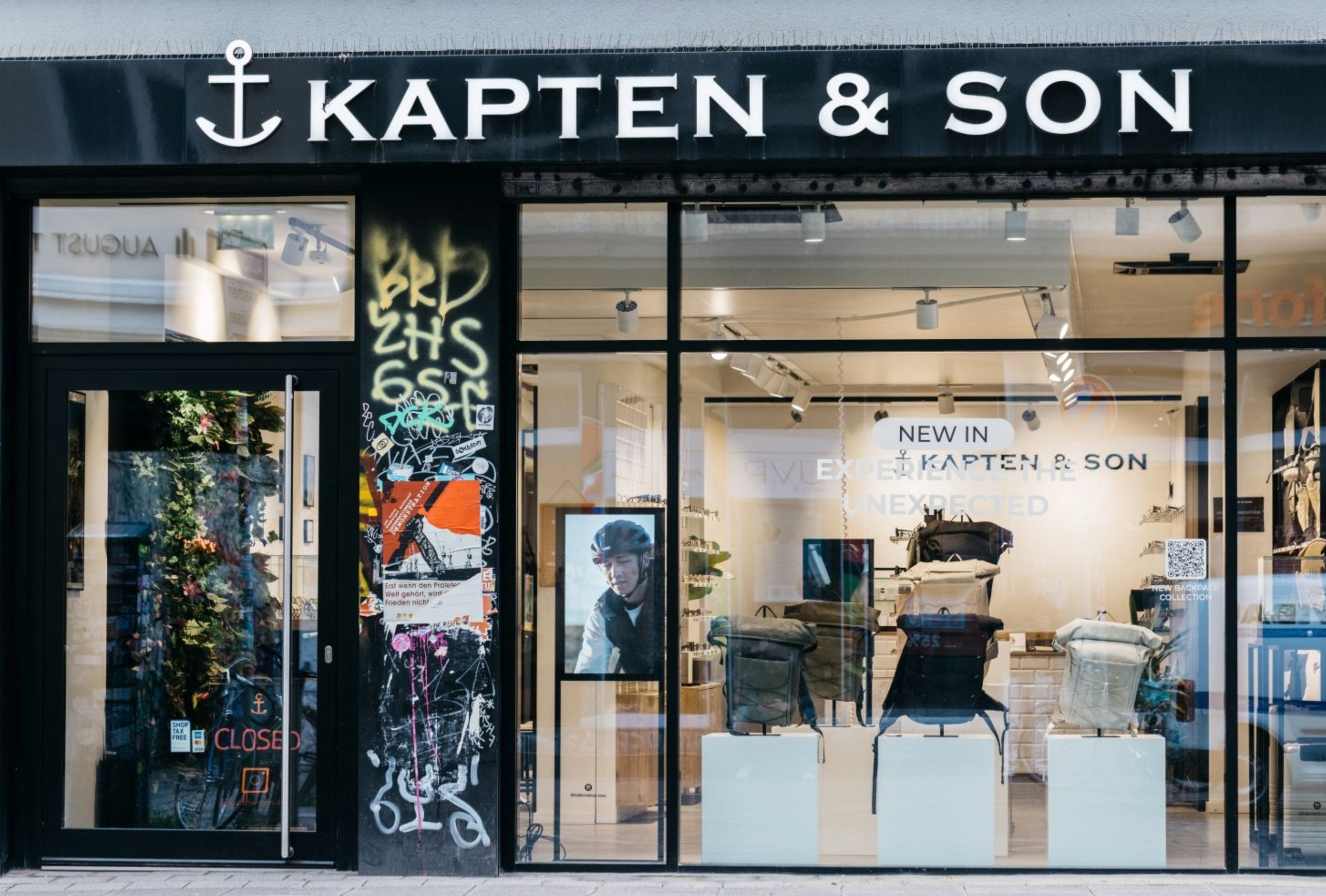 Kapten & Son Experience Store – Ladenfront mit beleuchtetem Logo, Rucksäcken, Taschen und Reisegepäck in den Schaufenstern