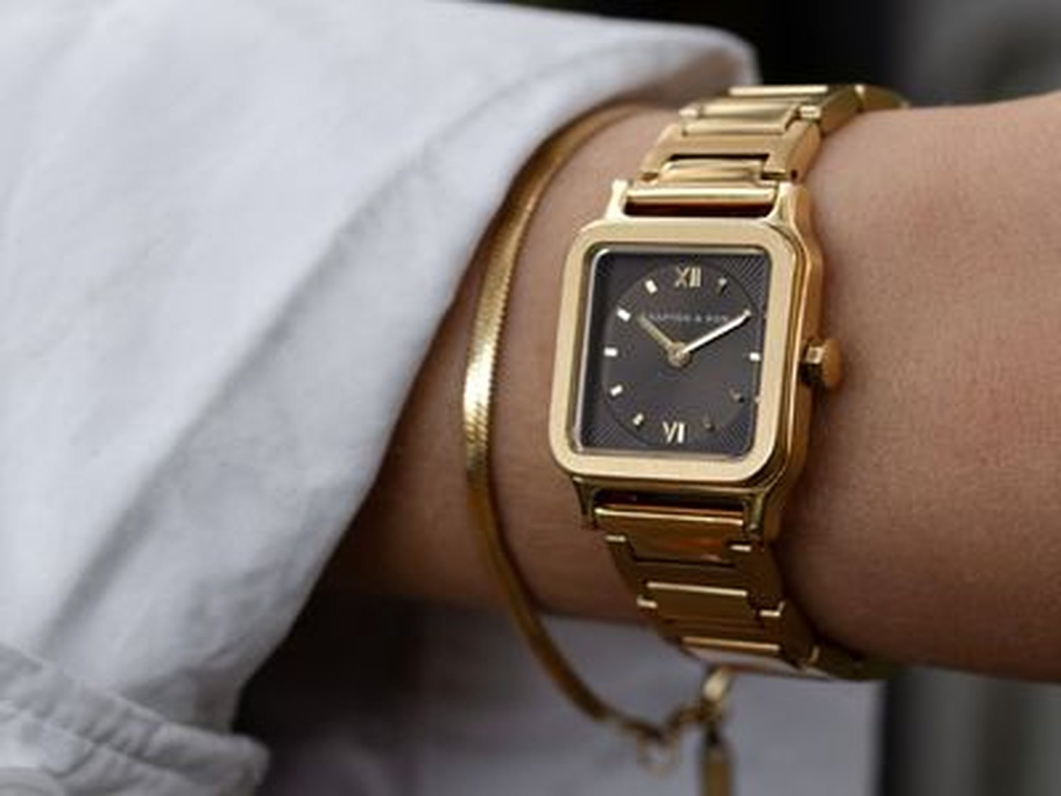 Gros plan d'un poignet portant une montre rectangulaire de Kapten & Son en or avec un cadran brun et des chiffres romains, associée à un bracelet doré.