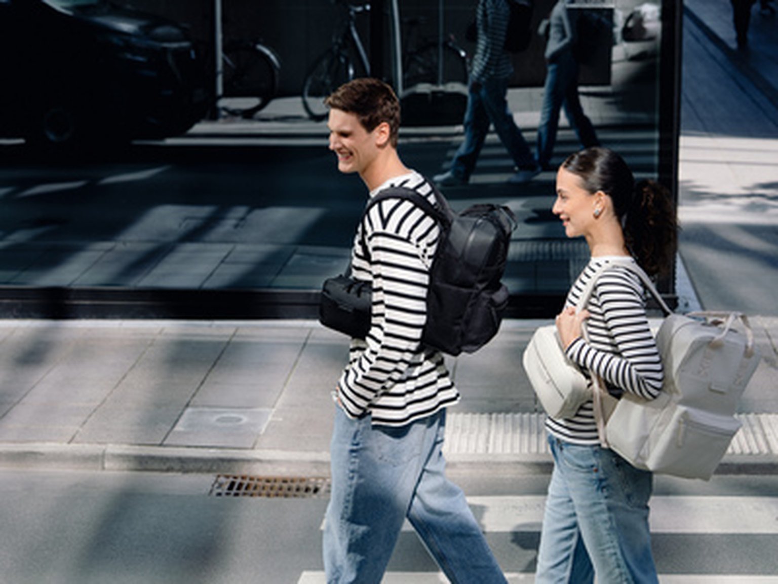 Deux personnes souriantes en chemises rayées traversent une rue. L'homme porte un sac à dos noir de Kapten & Son, la femme un sac à dos gris clair de Kapten & Son.