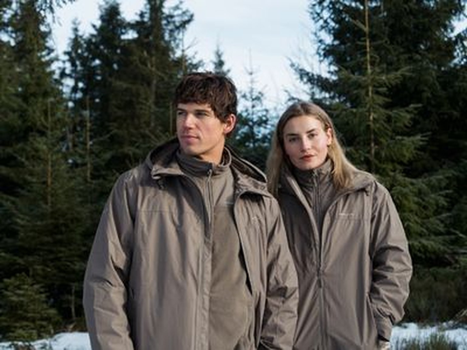 Un homme et une femme se tenant dans un paysage forestier enneigé, portant des vestes outdoor assorties de couleur taupe de Kapten & Son.