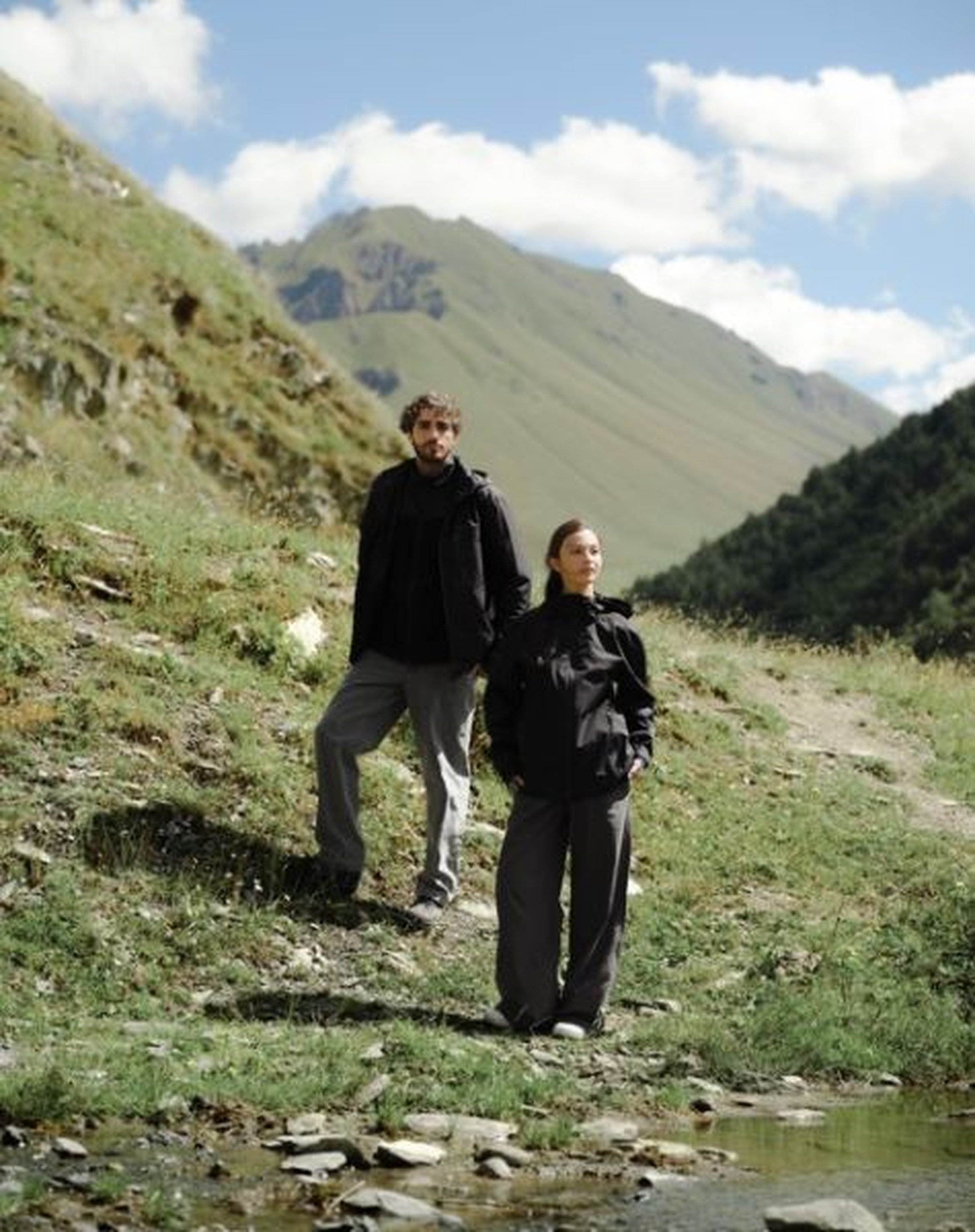 Een man en een vrouw staan op een wandelpad in een bergachtig landschap, gekleed in zwarte outdoorjassen van Kapten & Son en grijze broeken.