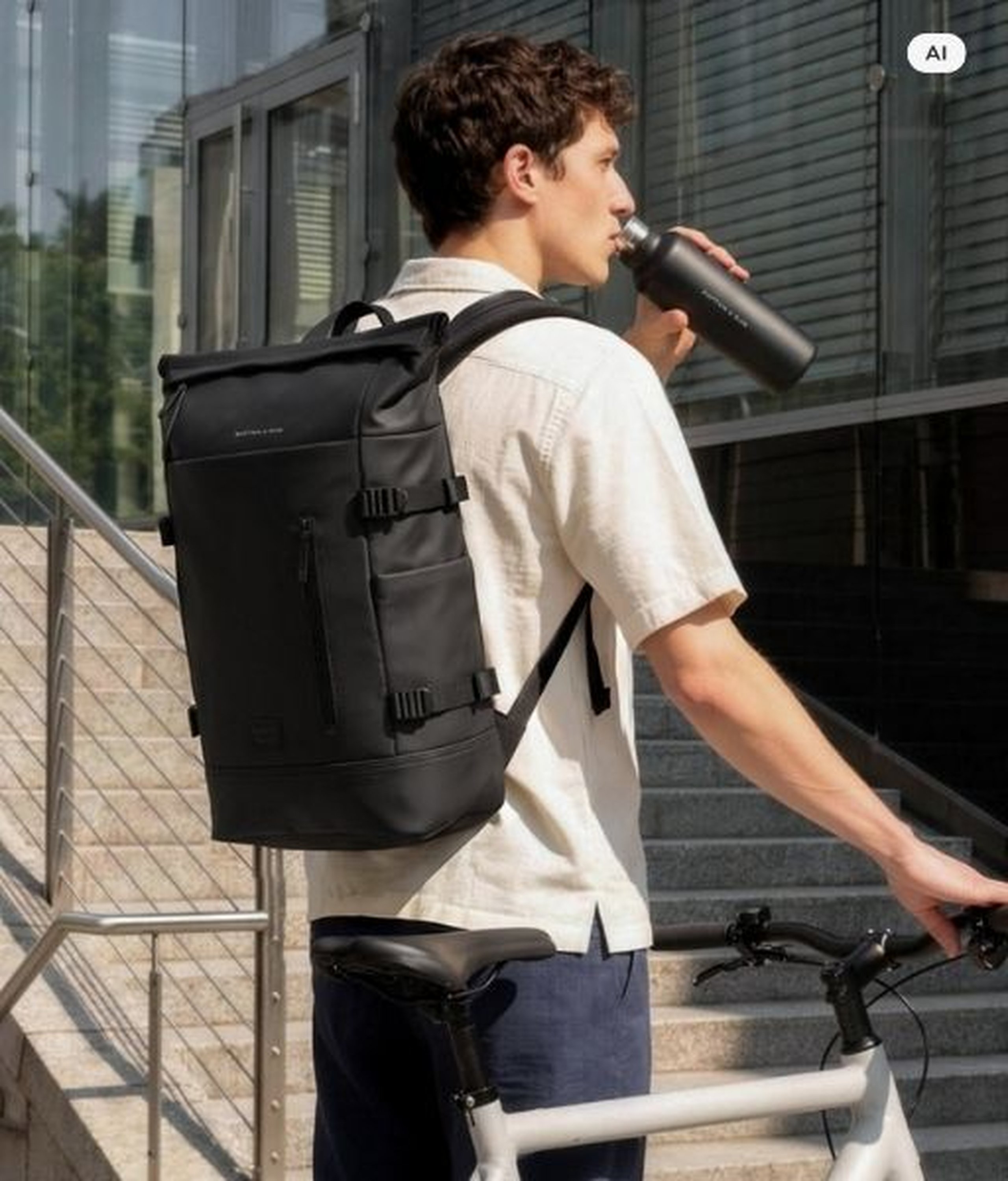 AI Generated Image: Een man met een zwarte Kapten & Son rolltop rugzak houdt een zwarte Kapten & Son drinkfles vast naast een witte fiets voor een modern gebouw.