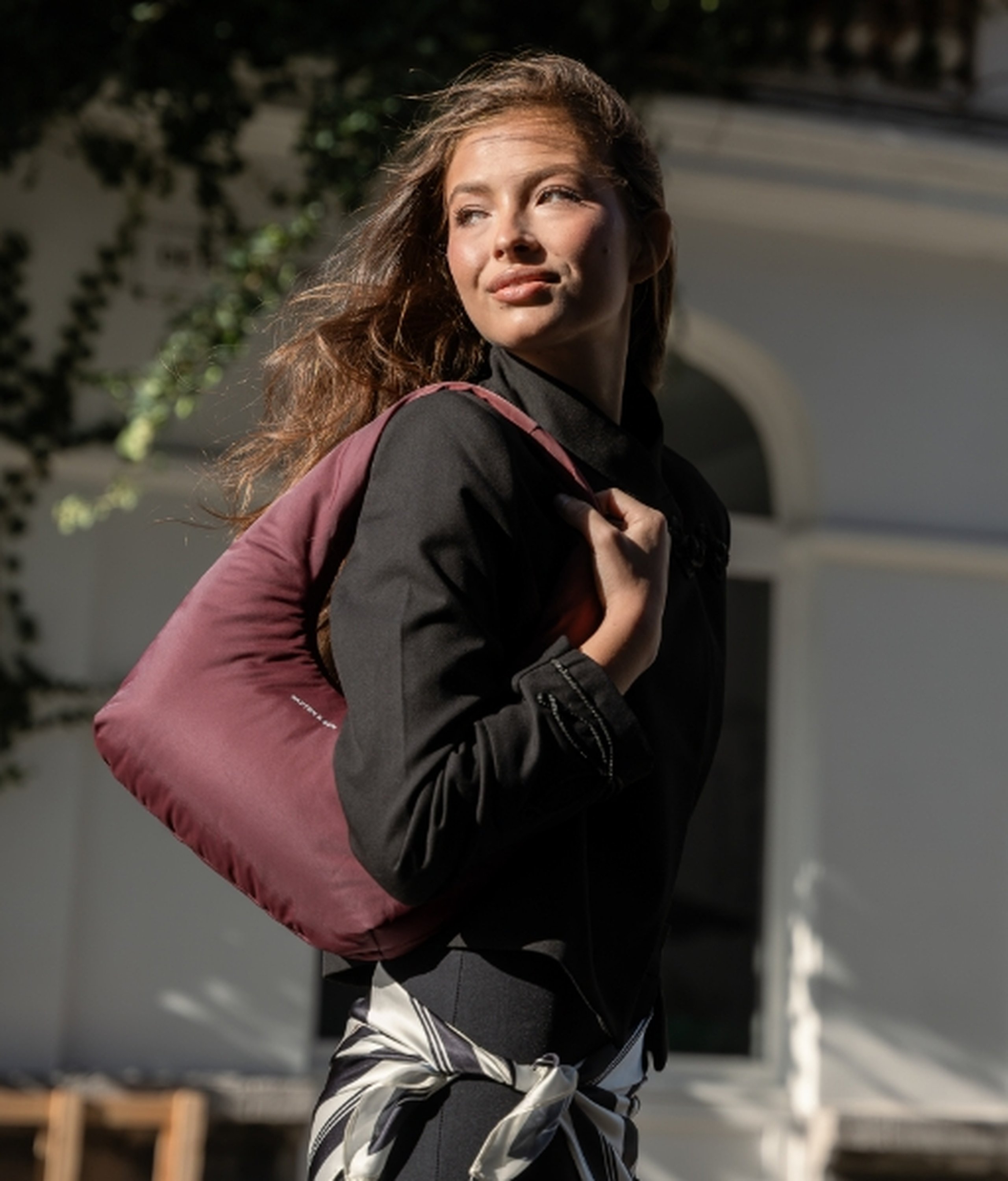 Junge Frau mit langen welligen Haaren blickt im Sonnenlicht nach oben, trägt eine schwarze Jacke und ein gemustertes Tuch um die Taille gebunden, hat eine große burgunderrote Kapten & Son Cloud-Schultertasche über der Schulter vor einem weißen Gebäude mit grünem Blattwerk.