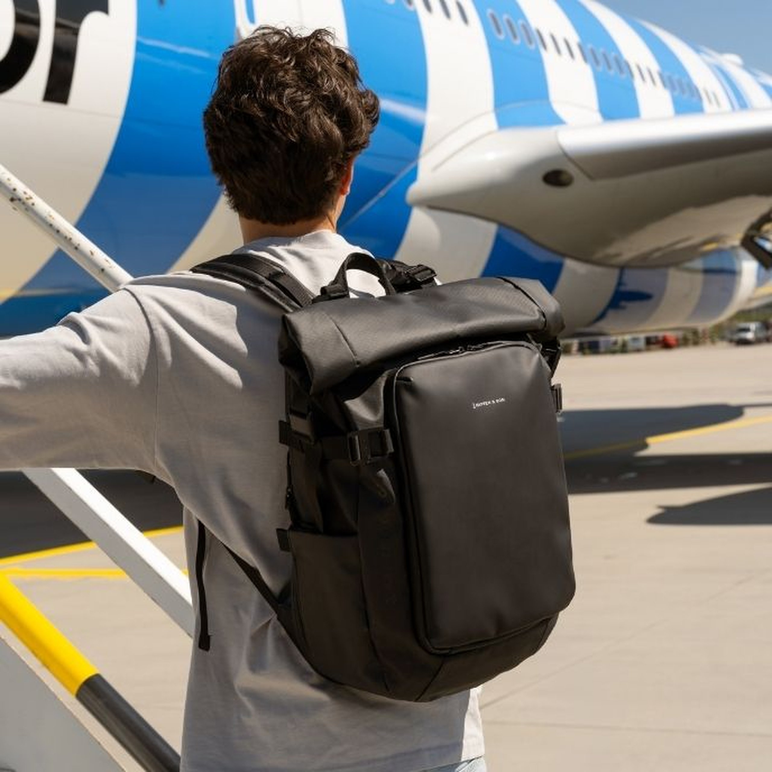 Junger Mann von hinten auf dem Flughafenvorfeld vor einem blau-weißen Flugzeug stehend, trägt ein hellgraues Langarmshirt und einen großen schwarzen Kapten & Son Roll-Top-Rucksack.