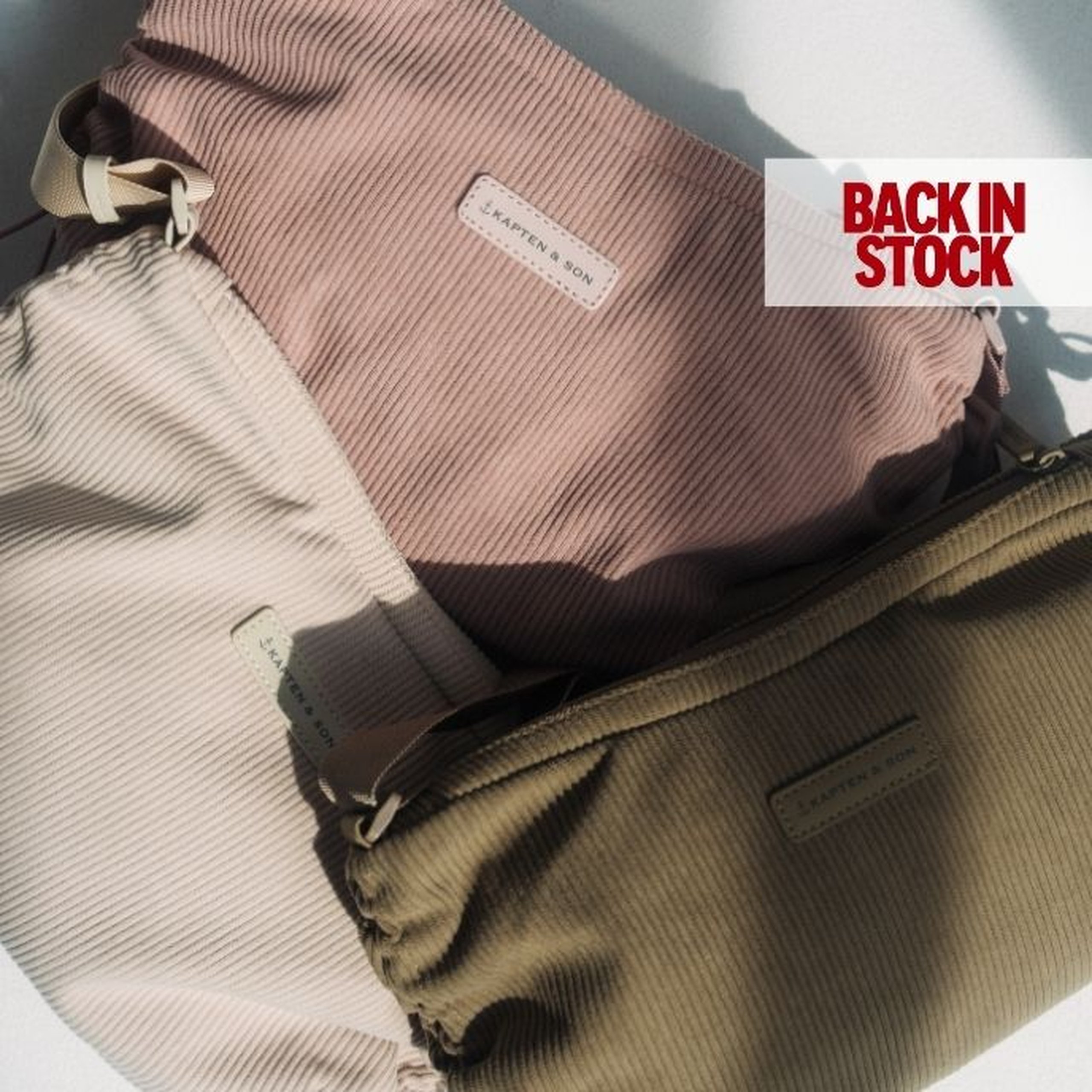 Tres bolsos de hombro de pana de Kapten & Son en crema, rosa polvorienta y verde oliva, superpuestos sobre una superficie clara bajo la luz del sol, con una etiqueta "BACK IN STOCK" en la esquina superior derecha.