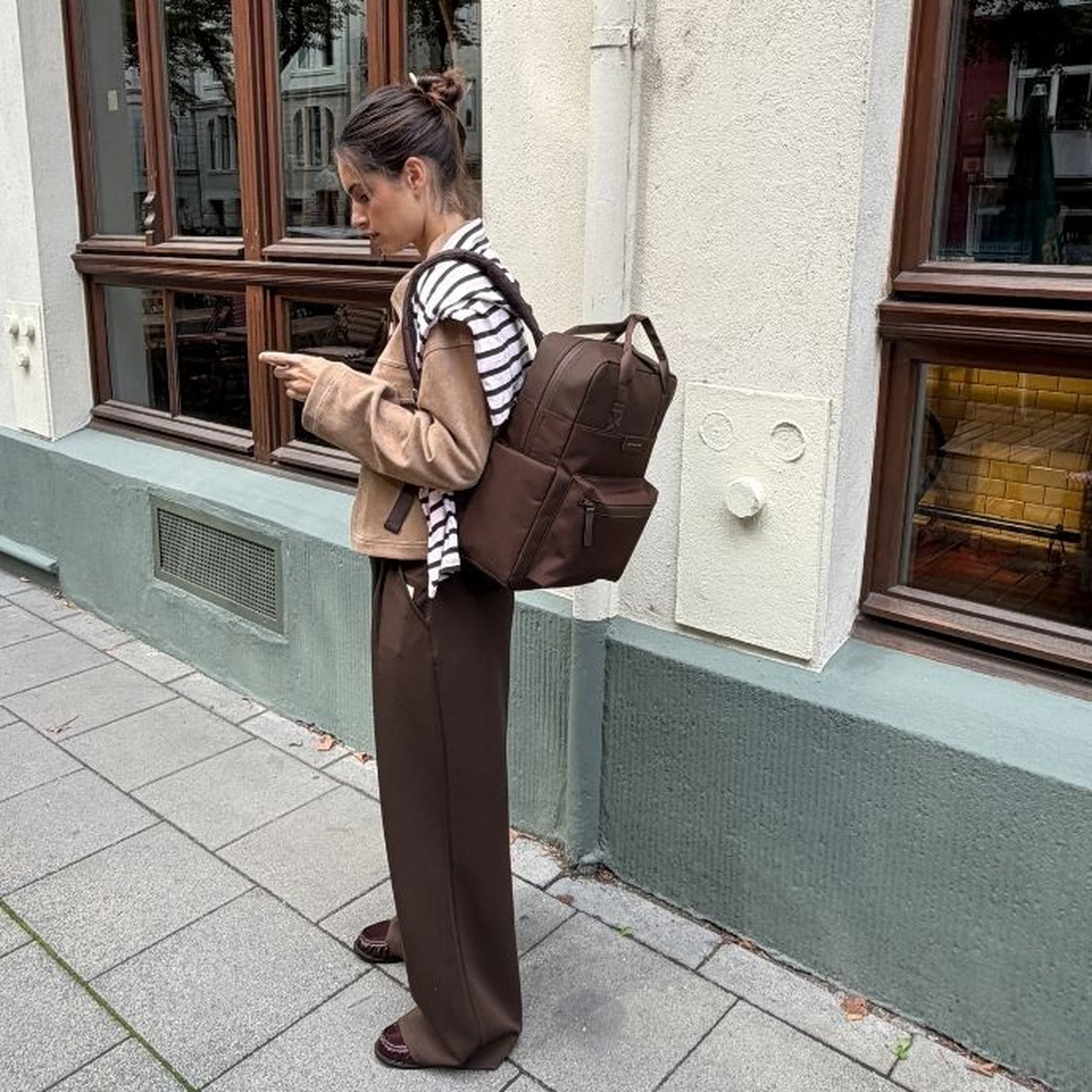 Jeune femme aux cheveux foncés en chignon debout sur un trottoir en ville en regardant son téléphone, portant une veste beige sur un top rayé noir et blanc, un pantalon large marron foncé et des mocassins marron foncé, avec un sac à dos Kapten & Son Bergen Pro couleur chocolat avec poignées supérieures et plusieurs poches zippées dans le dos.