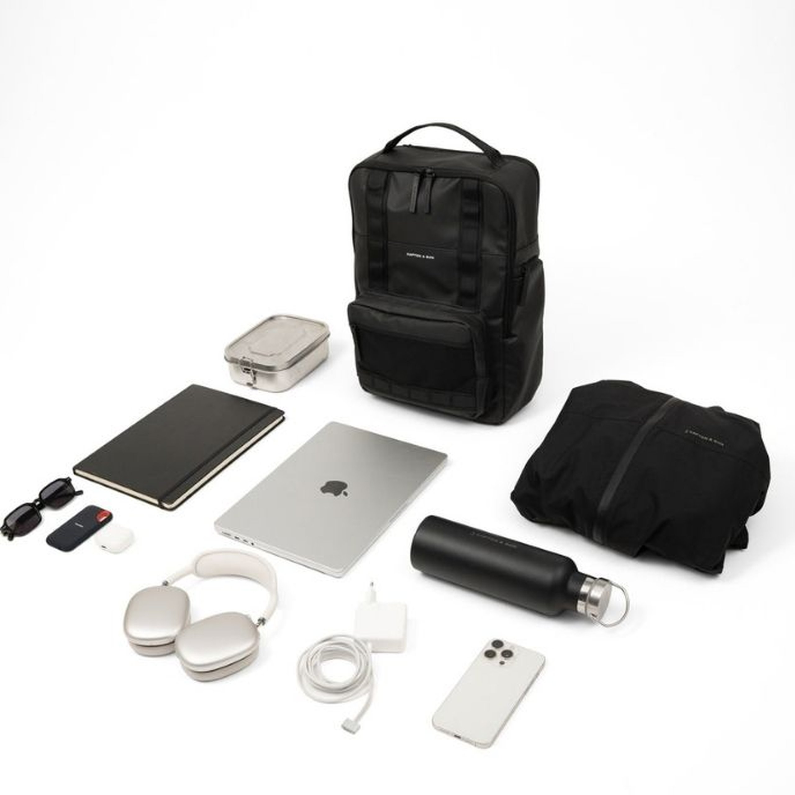 Flat Lay eines schwarzen Kapten & Son Bergen Ultra Rucksacks mit seinem Inhalt auf weißem Hintergrund: ein MacBook Laptop, weiße Over-Ear-Kopfhörer, ein schwarzes Notizbuch, eine Sonnenbrille, ein USB-Stick, ein Laptop-Ladekabel, ein Smartphone, eine schwarze Edelstahl-Trinkflasche, eine Metall-Lunchbox, eine gefaltete schwarze Kapten & Son Regenjacke und diverses Zubehör.
