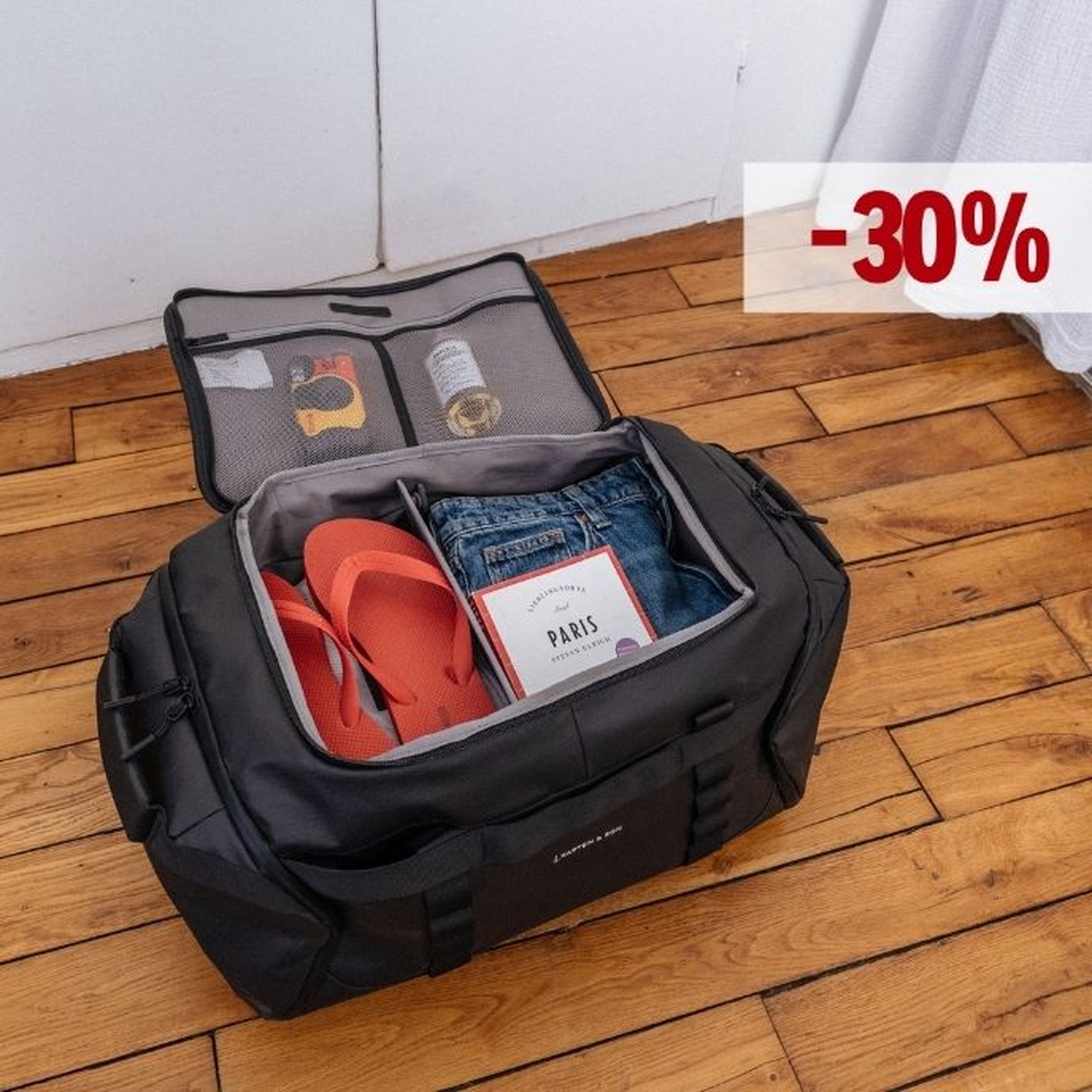 Geöffnete schwarze Kapten & Son Duffle Bag liegt auf einem Holzboden, gepackt mit roten Flip-Flops, Jeans, einem Paris-Reiseführer und Reiseaccessoires im Netzfach des Deckels, mit einem „-30%"-Rabatt-Label in der oberen rechten Ecke.