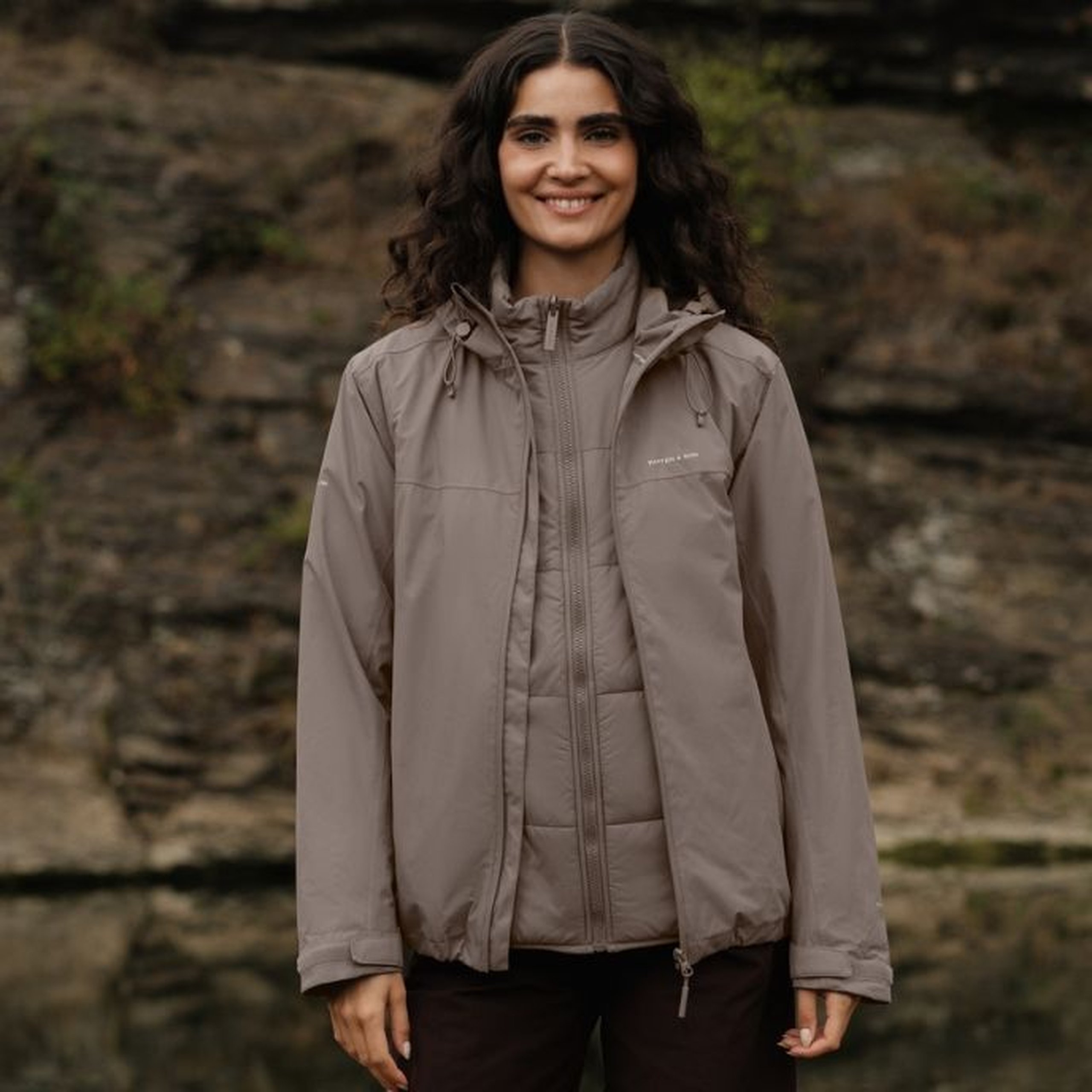Smiling young woman with long curly dark hair wearing a taupe Kapten & Son 3-in-1 puffer rain jacket with a high collar, chest logo and quilted inner layer visible, with dark brown trousers, standing in front of a rocky natural backdrop.