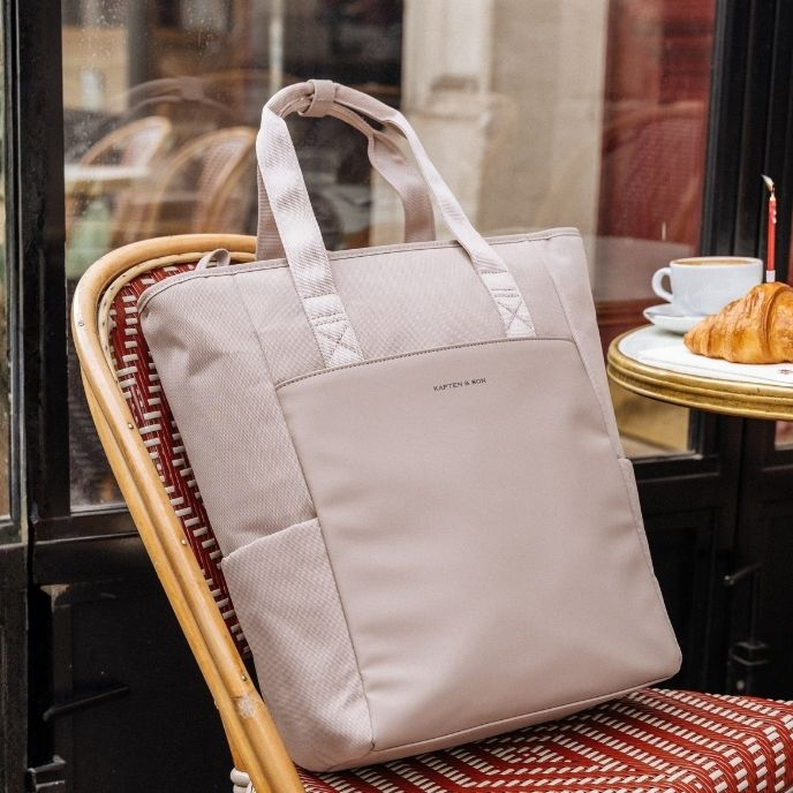 Large light pink Kapten & Son diaper tote bag with top handles and a structured front panel, placed on a red wicker café chair in a Parisian café setting, with a croissant and a coffee cup visible on a bistro table in the background.