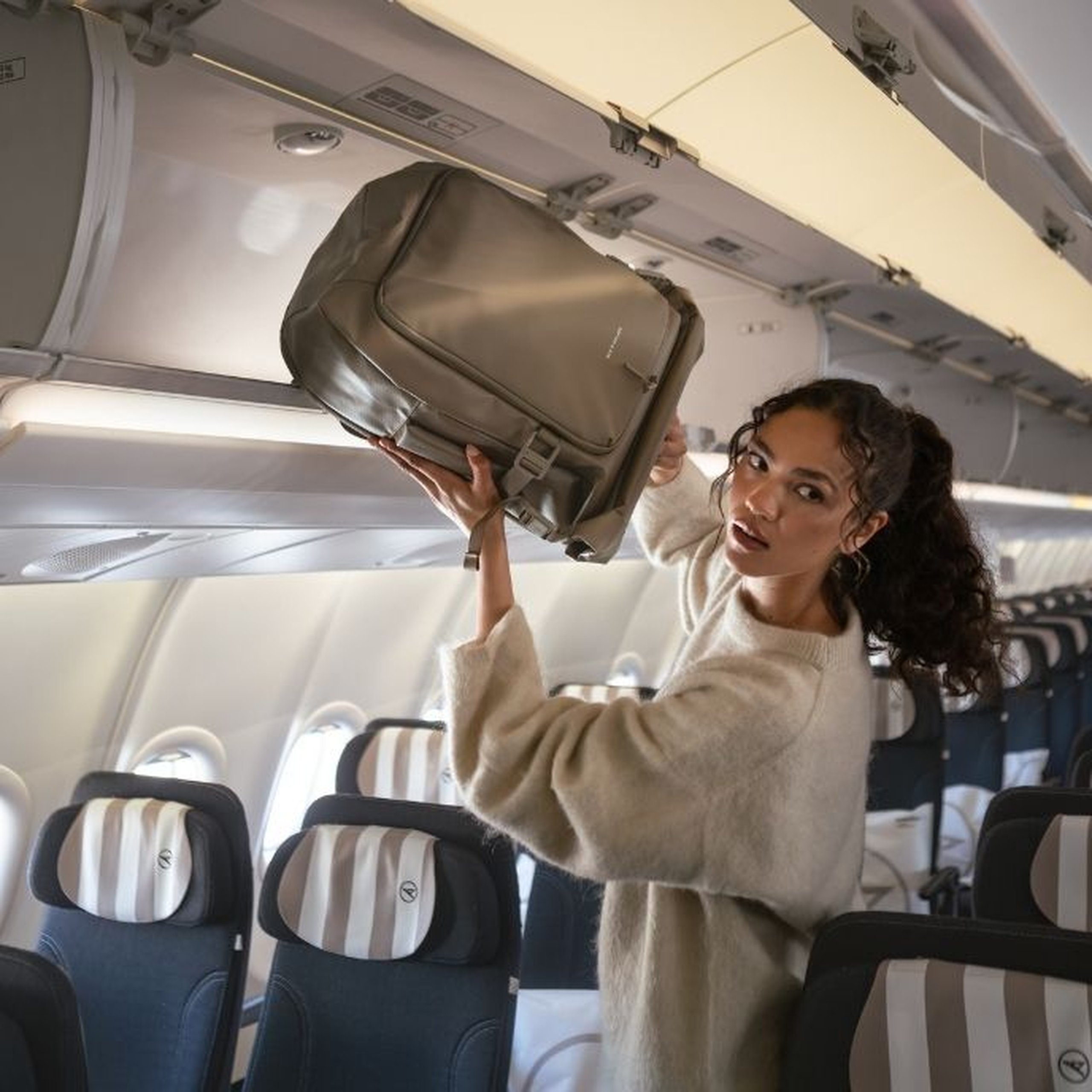 Eine Frau in einem cremefarbenen Pullover verstaut einen taupefarbenen Kapten & Son Rucksack im Gepäckfach eines Flugzeugs.