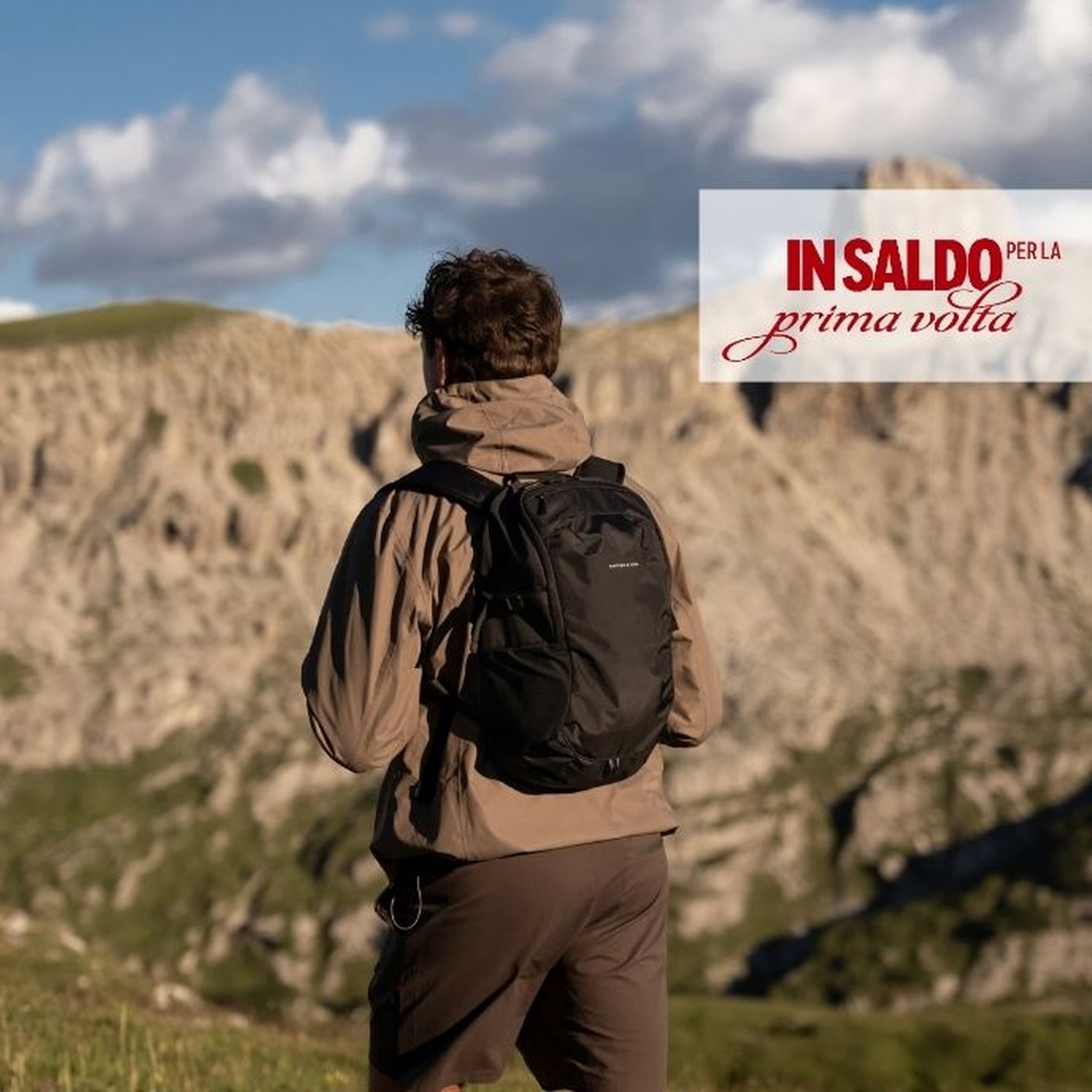 Uomo visto di spalle con una giacca beige con cappuccio e pantaloni scuri, con uno zaino daypack nero di Kapten & Son, in piedi davanti a un paesaggio montano suggestivo sotto un cielo nuvoloso. Un testo sovrapposto recita "first time ON SALE".