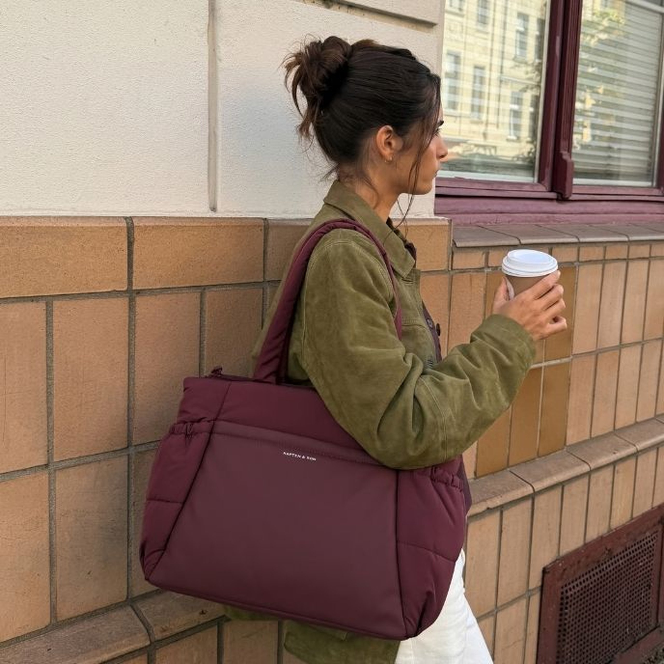 Jeune femme aux cheveux foncés en chignon appuyée contre un mur carrelé, portant une veste en daim vert olive et un pantalon blanc, tenant un café et portant un grand sac tote matelassé bordeaux Kapten & Son Hellvi Cloud sur l'épaule.