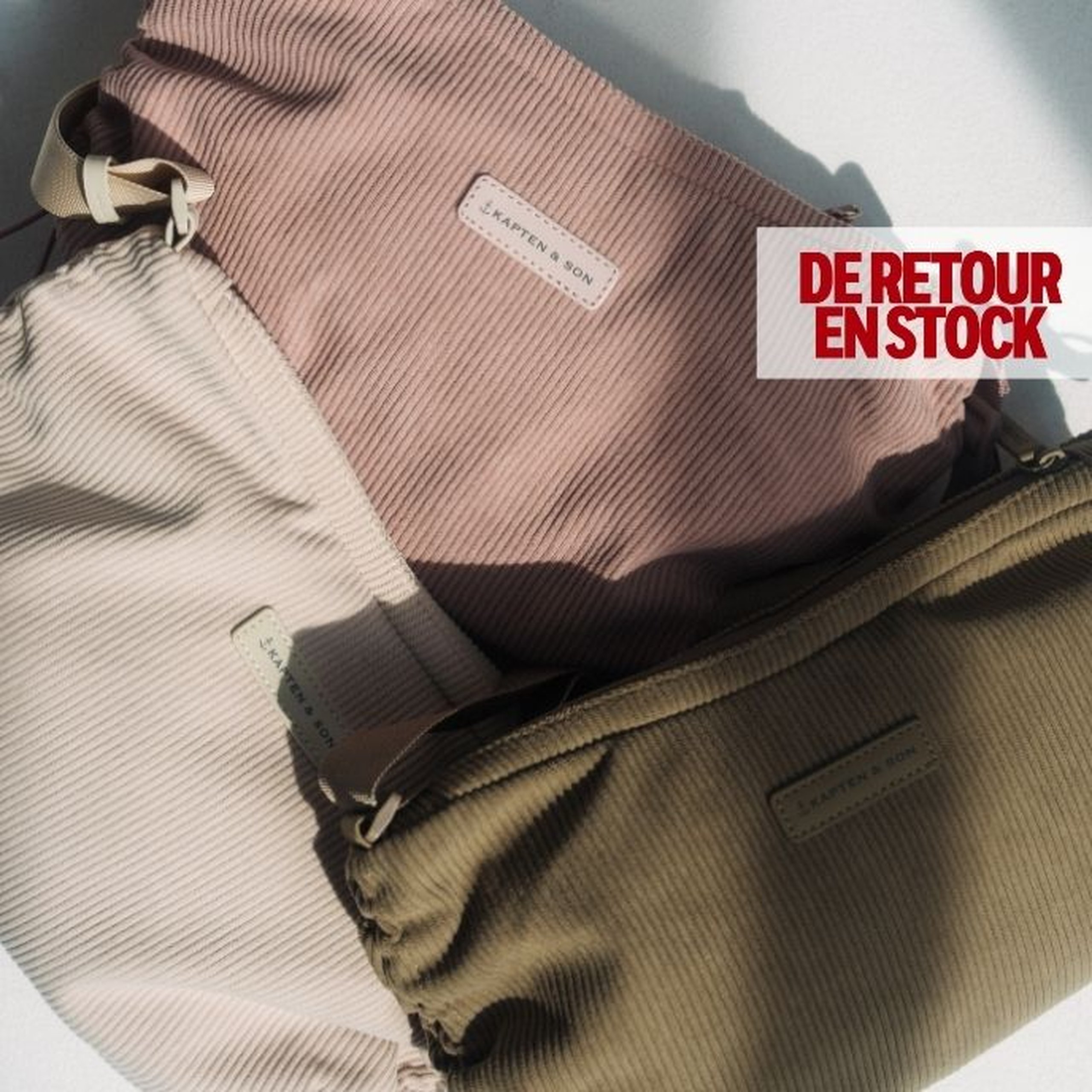 Trois sacs à bandoulière en velours côtelé Kapten & Son en crème, rose poudré et vert olive, se chevauchant sur une surface claire en plein soleil, avec une étiquette "BACK IN STOCK" dans le coin supérieur droit.