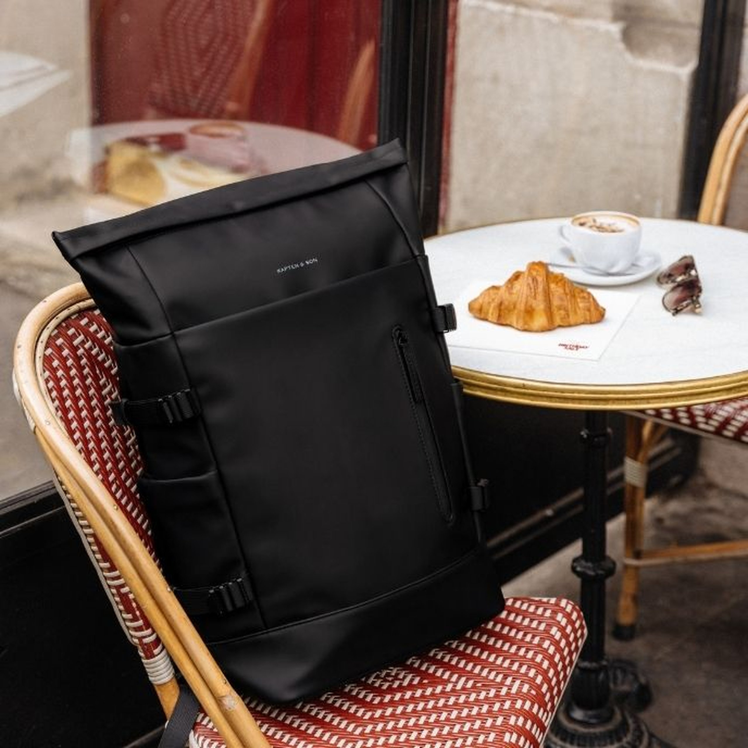 Black Kapten & Son roll-top backpack with side buckles and a front zip pocket placed on a red wicker café chair, next to a bistro table with a croissant, a cappuccino and sunglasses, in a Parisian café setting.