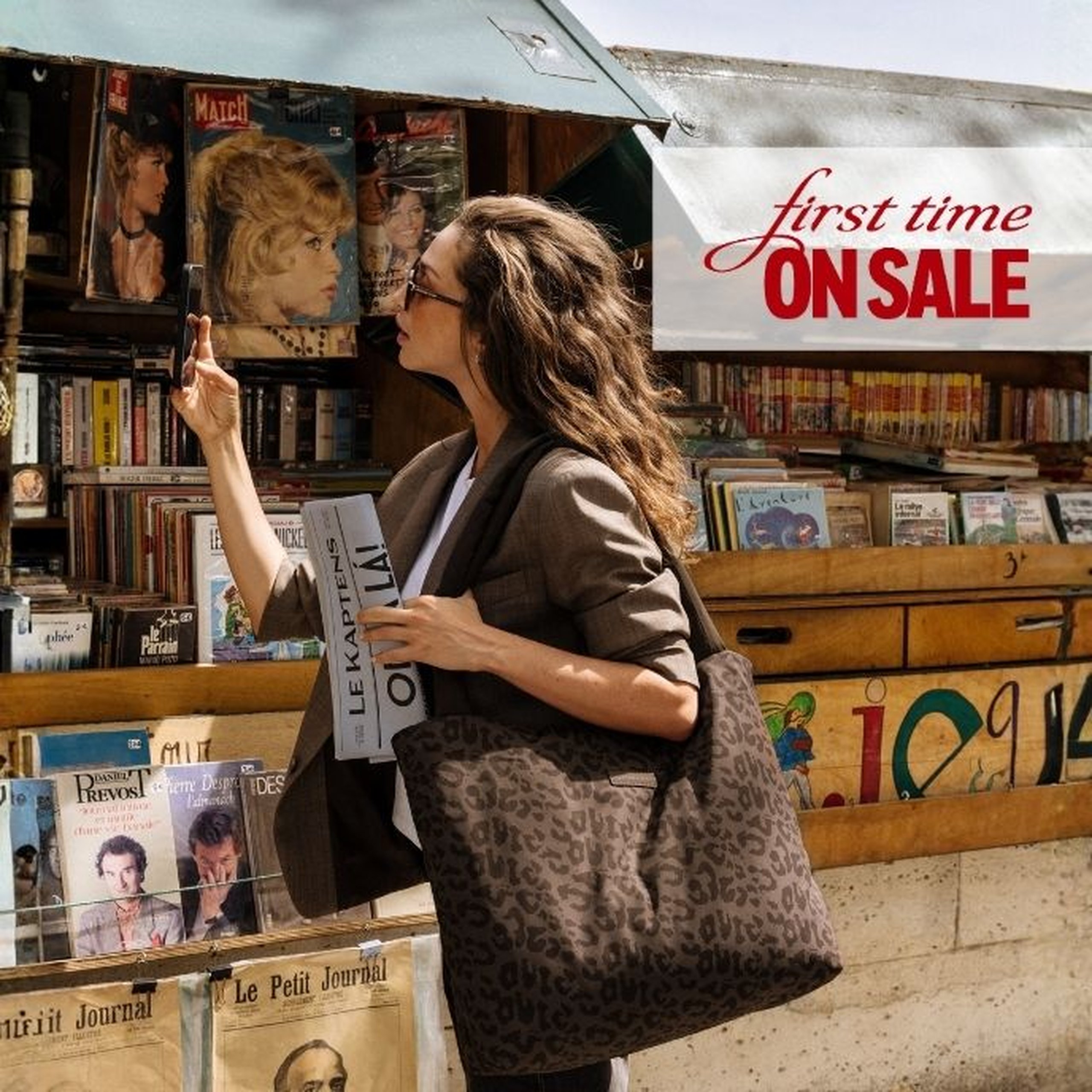 Woman with long wavy hair and glasses browsing books at a Parisian riverside bookstall, wearing a grey blazer and carrying a large dark brown leopard-print Kapten & Son tote bag, holding a "Le Kaptens – Oh là là!" newspaper. A text overlay reads "first time ON SALE".
