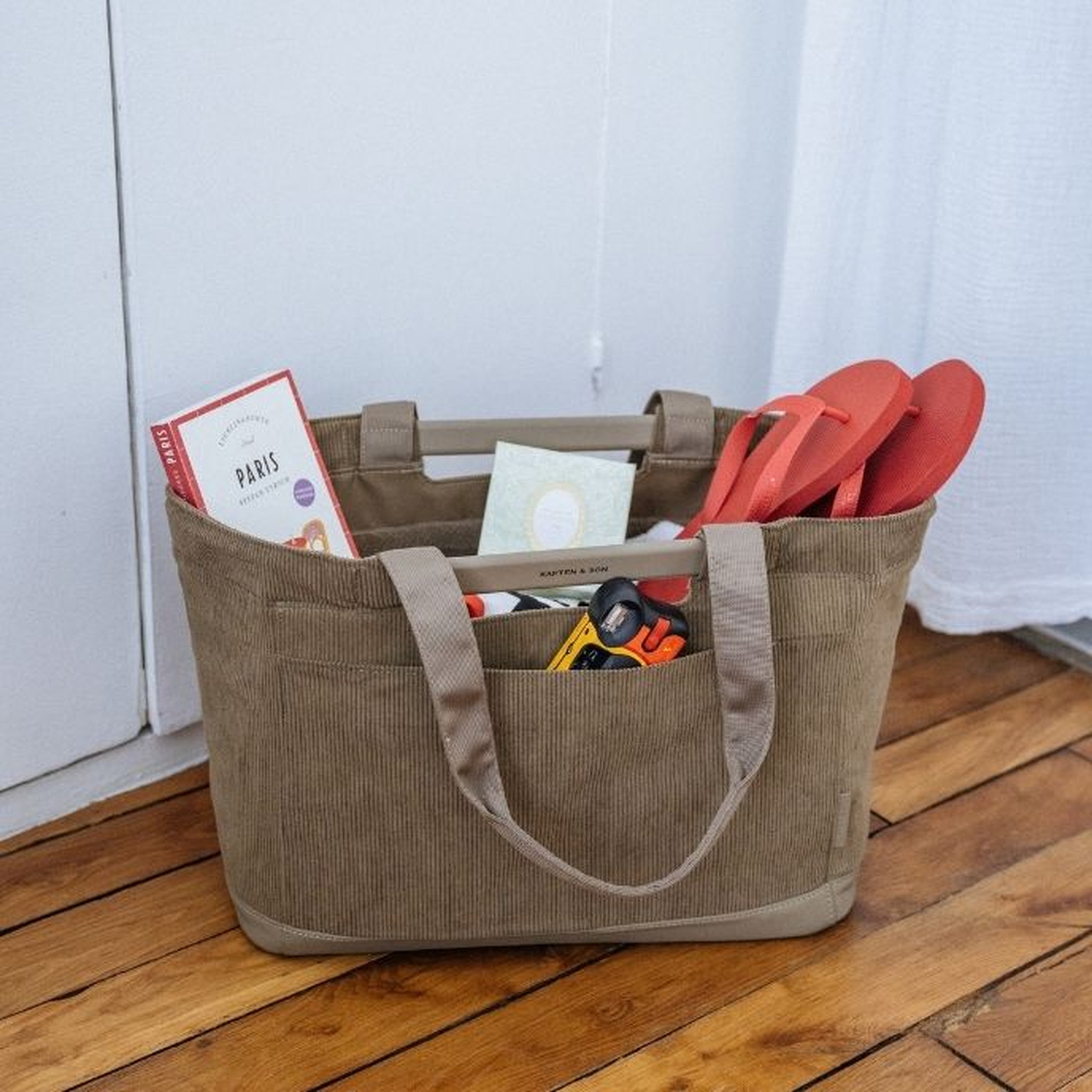 Grote stofkaki ribfluwelen Kapten & Son shopping tote staand op een houten vloer, gevuld met een reisgids van Parijs, een kaart, rode slippers en kleine accessoires in de voorvakken, geplaatst voor een witte deur.