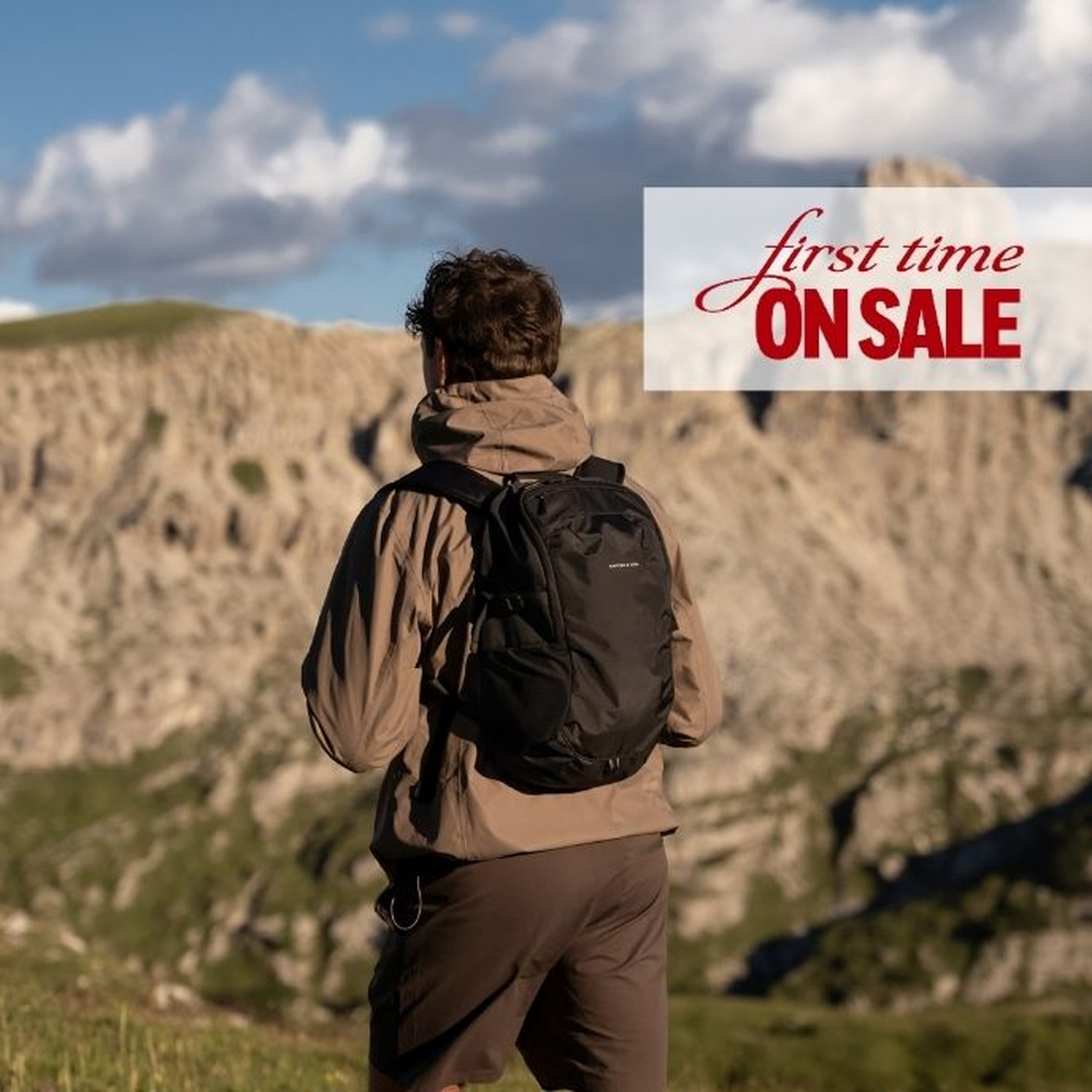 Man seen from behind wearing a beige hooded jacket and dark trousers, carrying a black Kapten & Son daypack, standing in front of a dramatic mountain landscape under a cloudy sky. A text overlay reads "first time ON SALE".