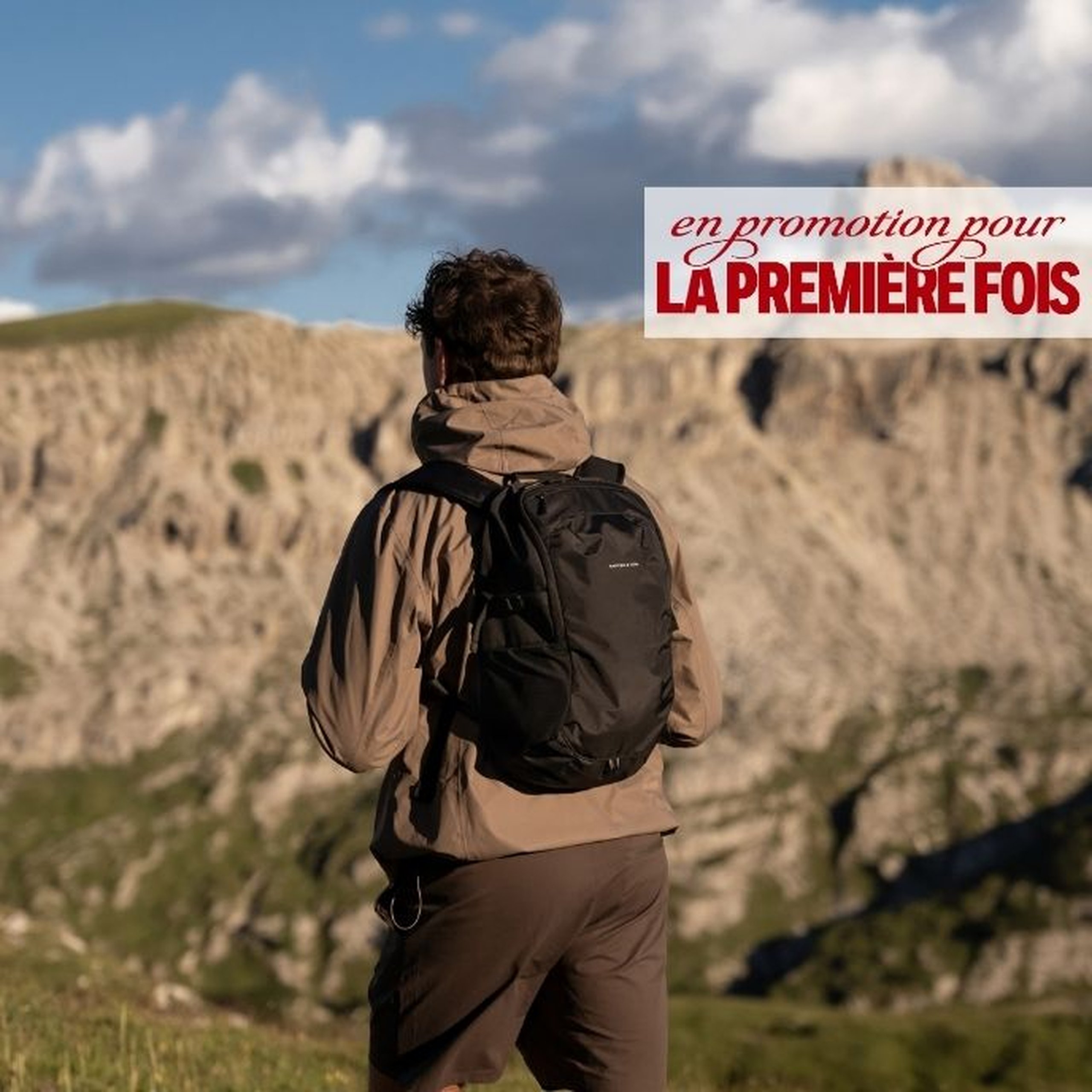 Homme vu de dos portant une veste à capuche beige et un pantalon sombre, avec un sac à dos daypack noir Kapten & Son, debout devant un paysage montagneux spectaculaire sous un ciel nuageux. Un texte superposé indique "first time ON SALE".