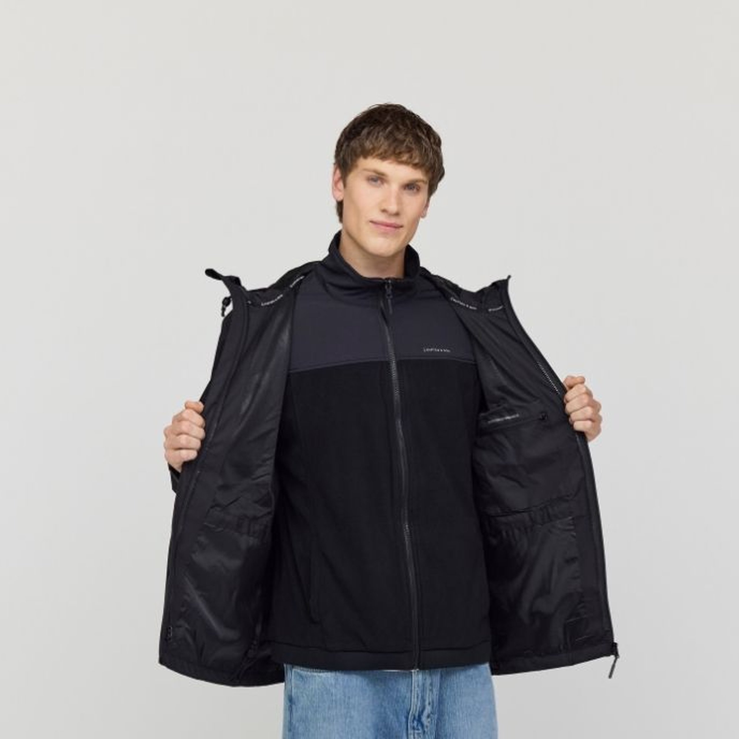 Young man holding open a black Kapten & Son 3-in-1 rain jacket to reveal the inner fleece layer and lining, wearing light blue jeans, against a light grey background.