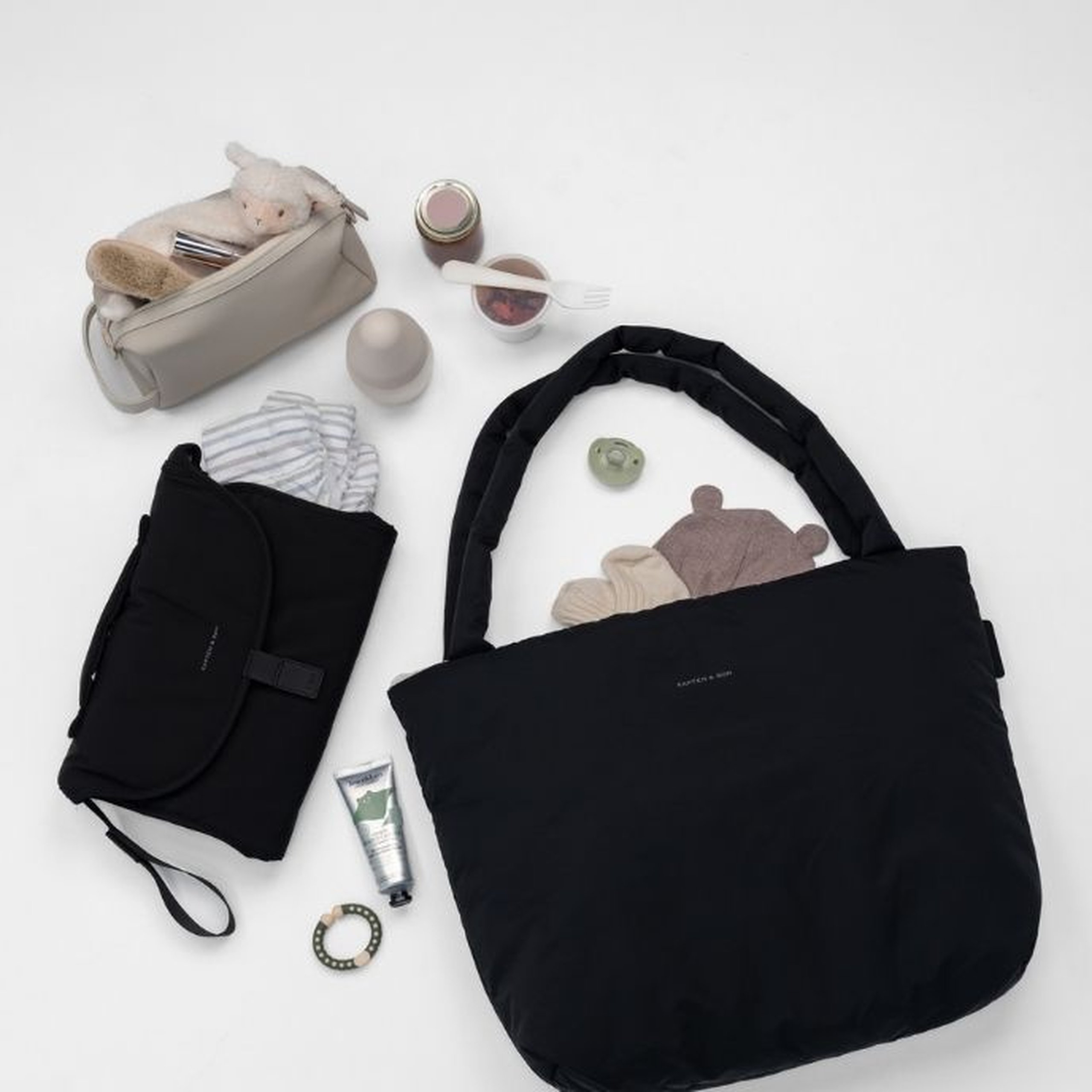Flat Lay einer schwarzen Kapten & Son Norva Wickeltasche mit gepolsterten Tragegriffen, umgeben von ihrem Inhalt auf einer weißen Fläche: eine schwarze faltbare Wickelunterlage mit Kapten & Son Logo, ein hellgrauer Kulturbeutel mit einem Kuscheltier und Kamm, Babynahrungsgläschen, ein Schnuller, ein Beißring, eine Handcreme-Tube, ein gestreifter Baby-Body und ein kleines Bären-Kuscheltier, das aus der Tasche herausschaut.