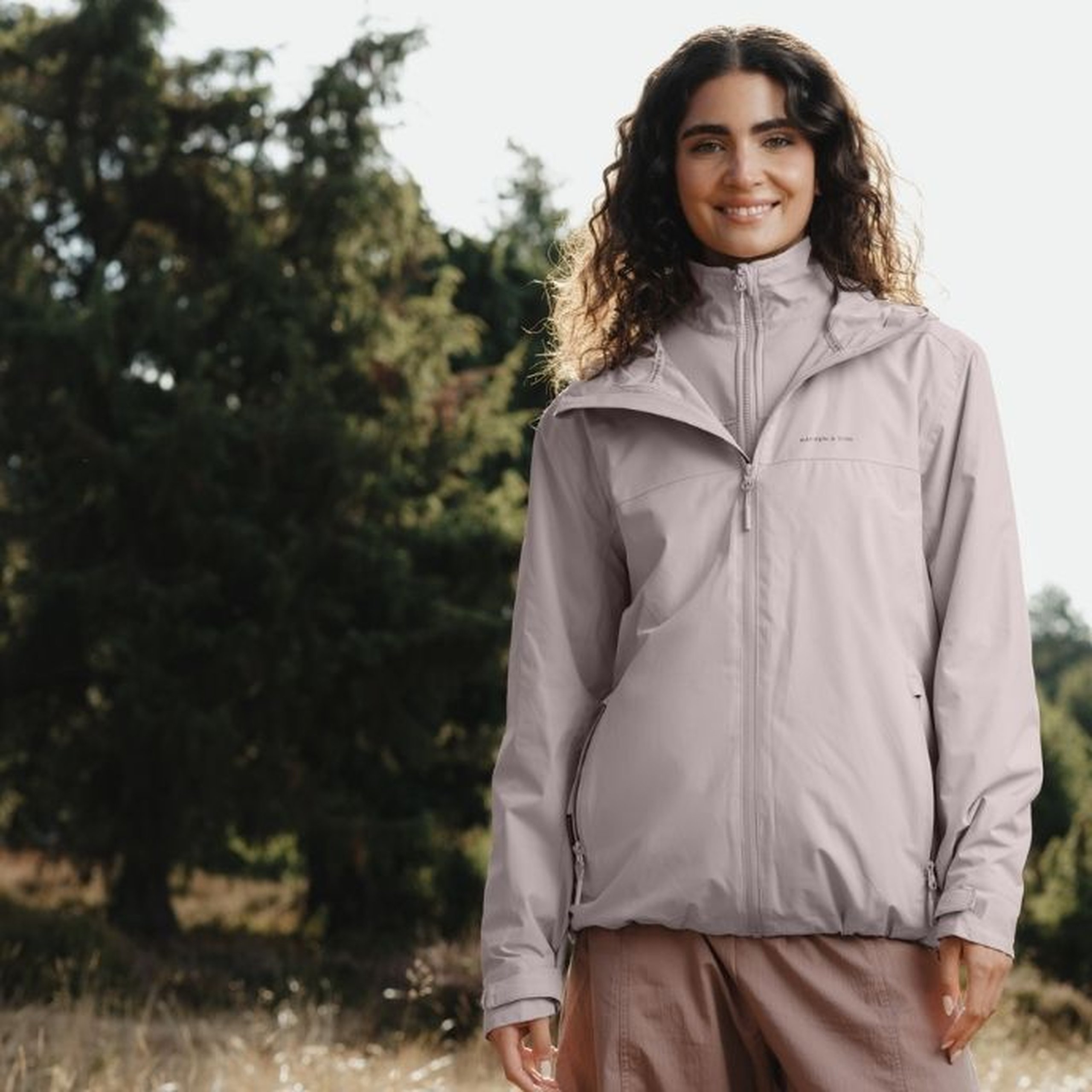 Smiling young woman with long curly hair wearing a zipped-up muted rose Kapten & Son rain jacket with a high collar, chest logo and side zip pockets, with taupe wide-leg trousers, standing in a sunny meadow with pine trees in the background.
