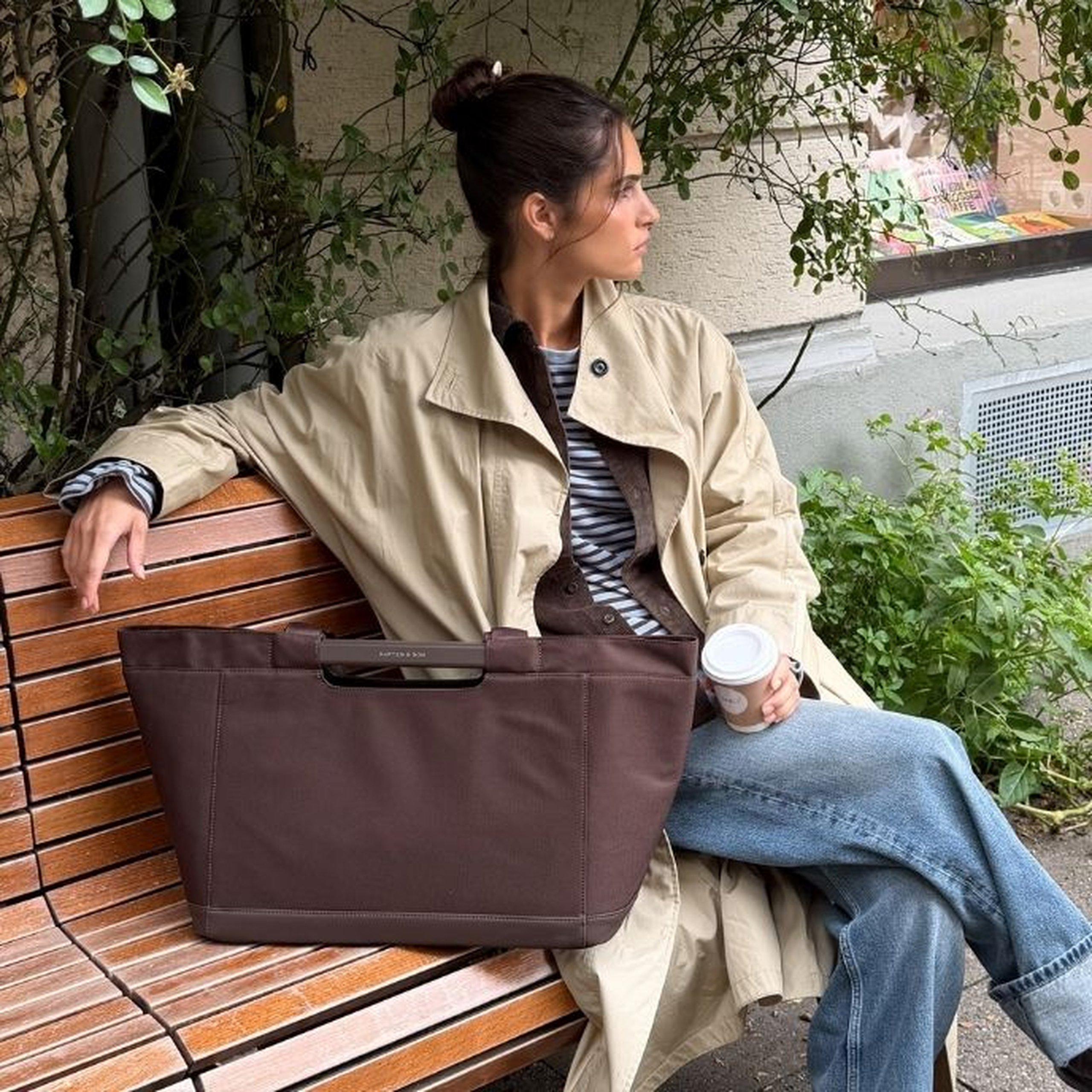 Jeune femme aux cheveux foncés en chignon assise sur un banc en bois à l'extérieur, portant un trench-coat beige sur un top rayé et un jean large bleu clair, tenant un café et posant un grand sac tote Kapten & Son Vaasa Shopping Basket couleur chocolat sur ses genoux.