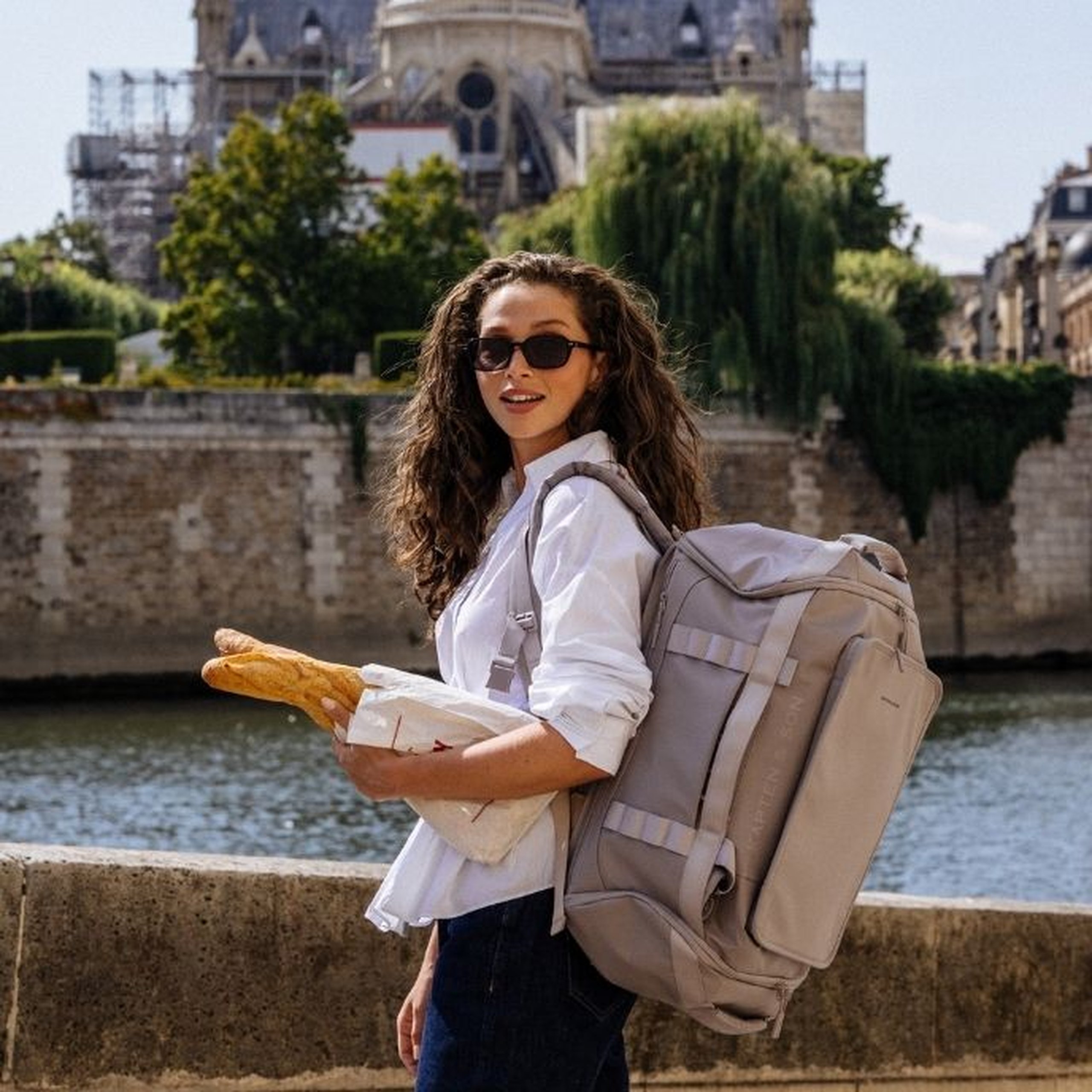 Junge Frau mit langen lockigen Haaren und Sonnenbrille trägt ein weißes Hemd und dunkle Jeans, hat einen großen hellgrauen Kapten & Son Duffle-Rucksack auf dem Rücken und hält ein Baguette in der Hand, steht auf einer Brücke an der Seine in Paris mit der Kathedrale Notre-Dame im Hintergrund.