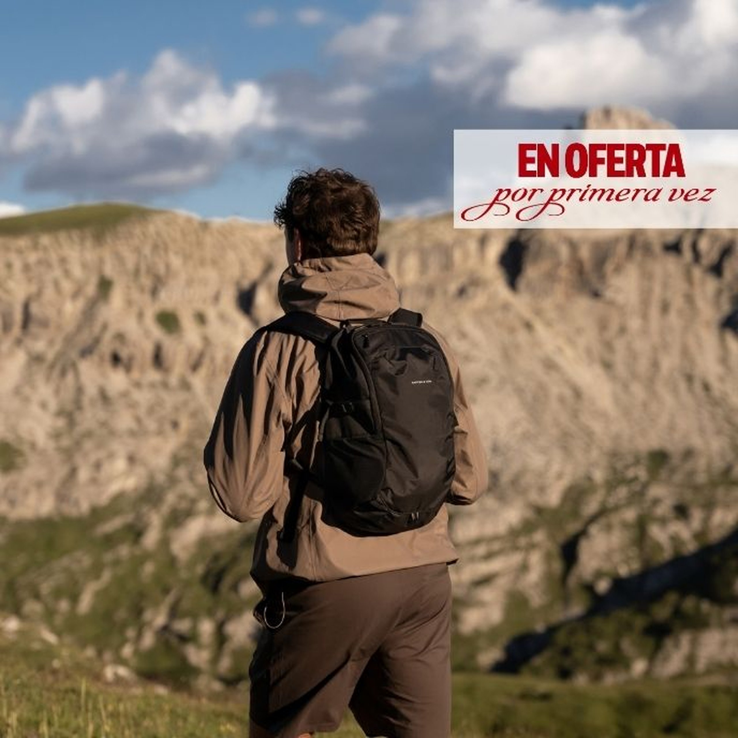 Hombre visto de espaldas con una chaqueta beis con capucha y pantalones oscuros, llevando una mochila daypack negra de Kapten & Son, de pie frente a un impresionante paisaje montañoso bajo un cielo nublado. Un texto superpuesto indica "first time ON SALE".