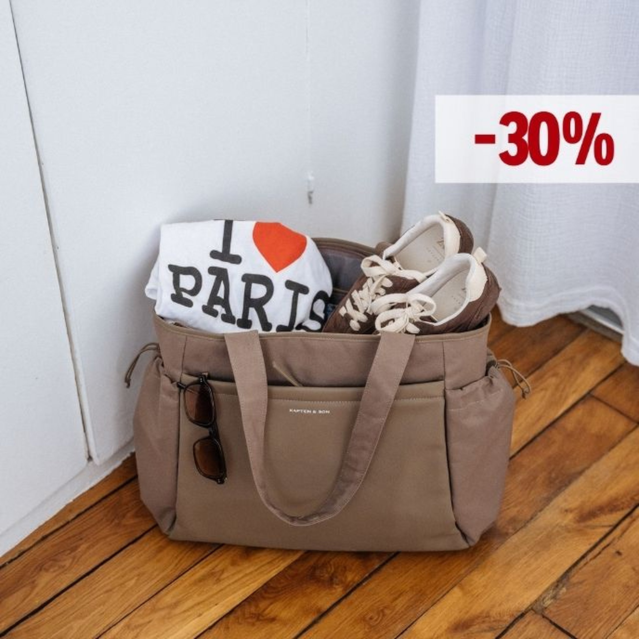 Gran bolso tote de Kapten & Son en color taupe sobre un suelo de madera, con una camiseta "I Love Paris" y un par de zapatillas marrones, con unas gafas de sol en el bolsillo lateral y una etiqueta de descuento "-30%" en la esquina superior derecha.
