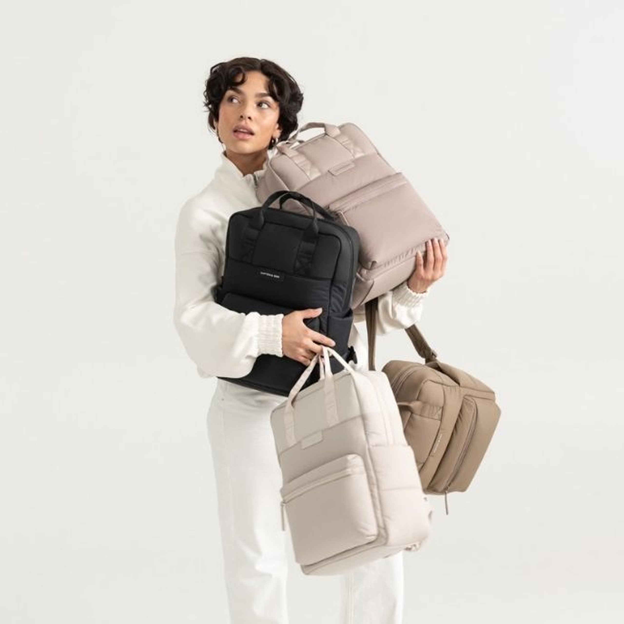 Junge Frau mit kurzem lockigem dunklem Haar in einem weißen Trainingsanzug hält mehrere Kapten & Son Taschen gleichzeitig vor einem hellgrauen Hintergrund: einen großen helltaupefarbenen gesteppten Bergen Cloud Rucksack über dem Kopf, eine schwarze Charles Laptoptasche im Arm, einen hellbeigen Bergen Cloud Rucksack vor sich und eine taupefarbene gesteppte Tasche am Arm hängend.