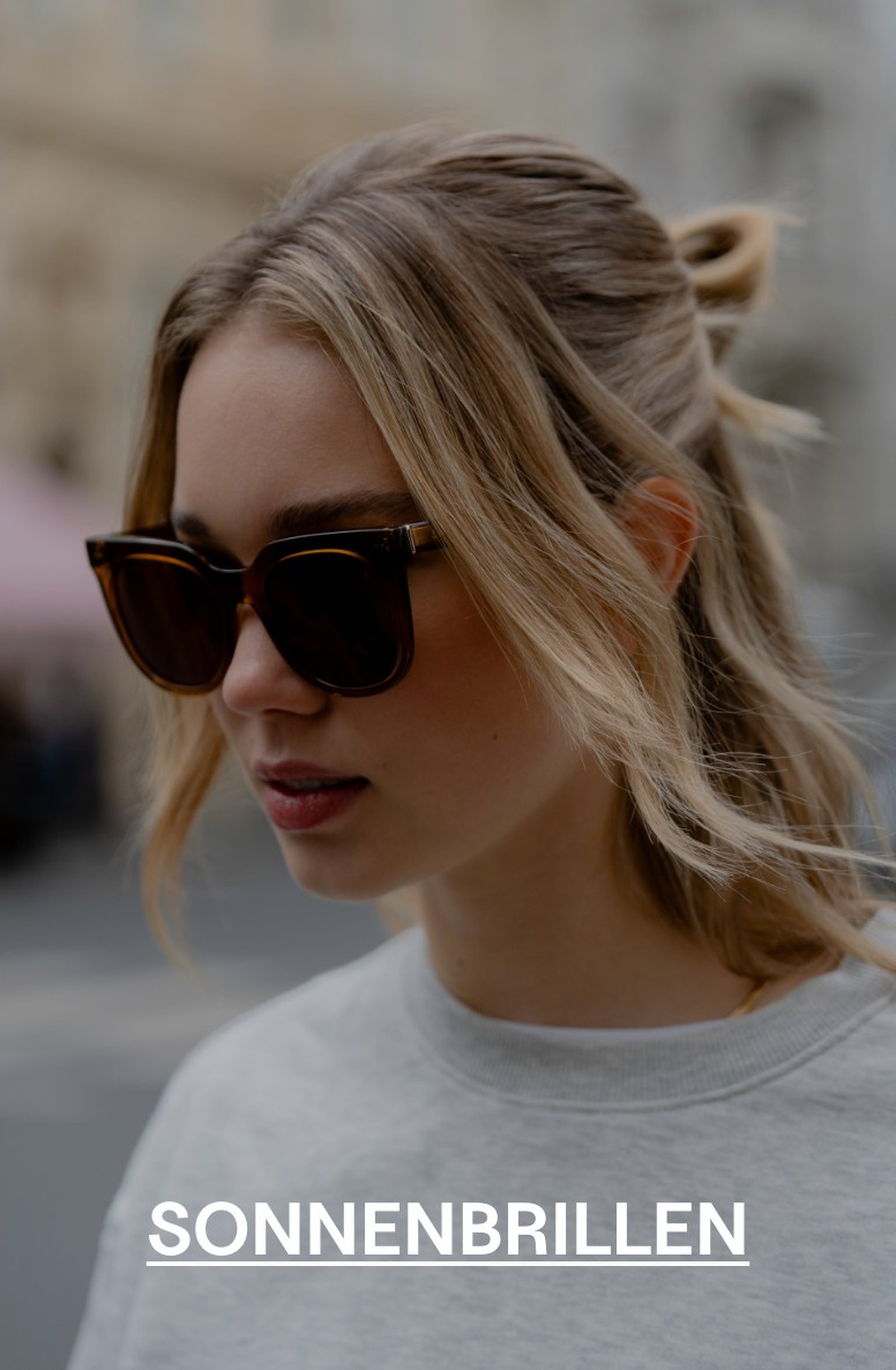 Frau mit blonden Haaren trägt eine Schildpatt-Sonnenbrille von Kapten & Son und ein hellgraues Sweatshirt, Blick nach unten gerichtet – Texteinblendung: „SONNENBRILLEN"