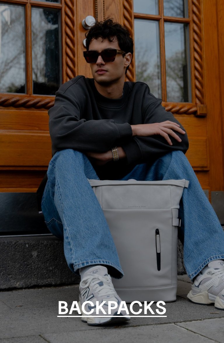 Man sitting on steps in front of a wooden door, wearing sunglasses and a grey sweatshirt, with a grey Kapten & Son backpack between his legs – text overlay: "BACKPACKS"